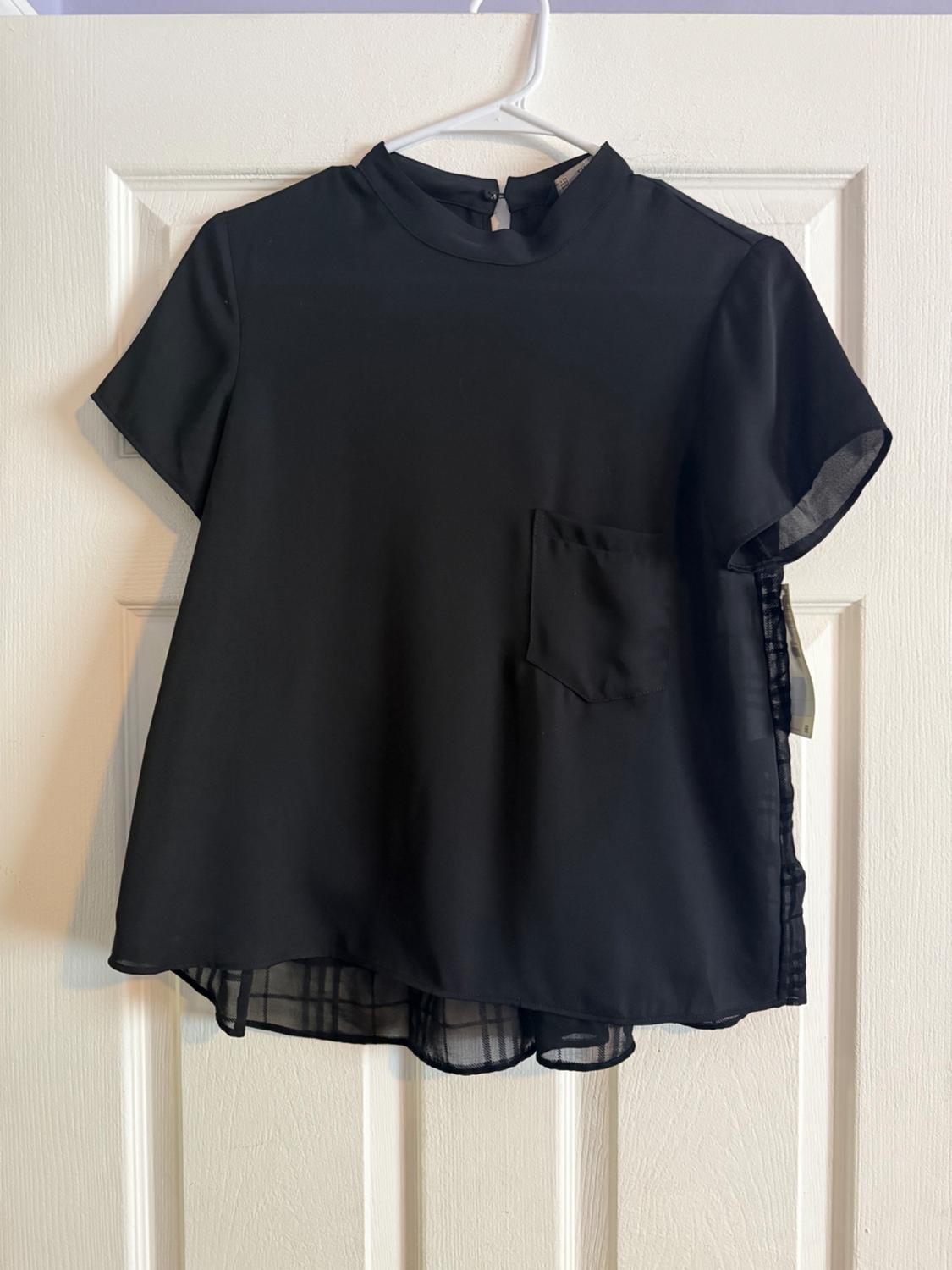 Black short-sleeved blouse with a high neck and front pocket, hanging on a white hanger over a white door.