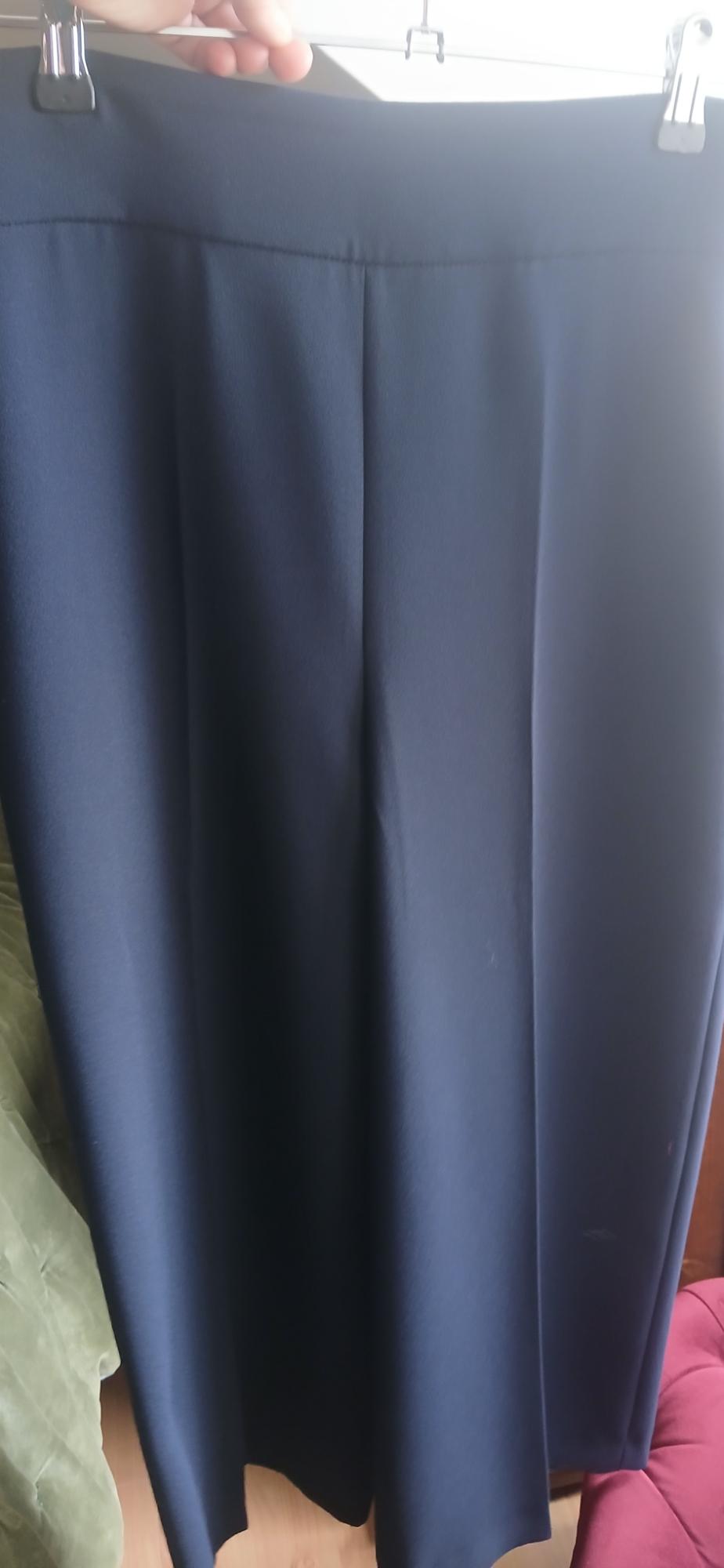Dark blue skirt hanging on a hanger, with soft pleats. Background with green and pink armchair. Natural light from the right.