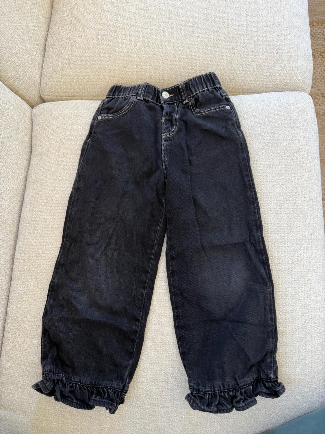Black denim pants with an elastic waistband and button, with ruffled cuffs, placed on a light beige sofa.