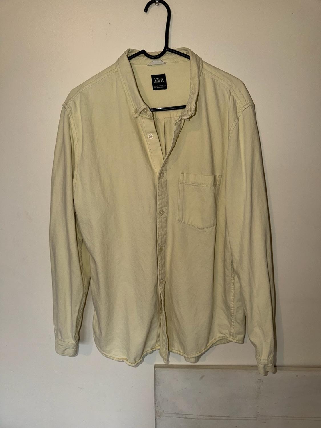 Light yellow long-sleeved shirt with a front pocket, hanging on a black hanger against a wall. Text on the label: ZARA.