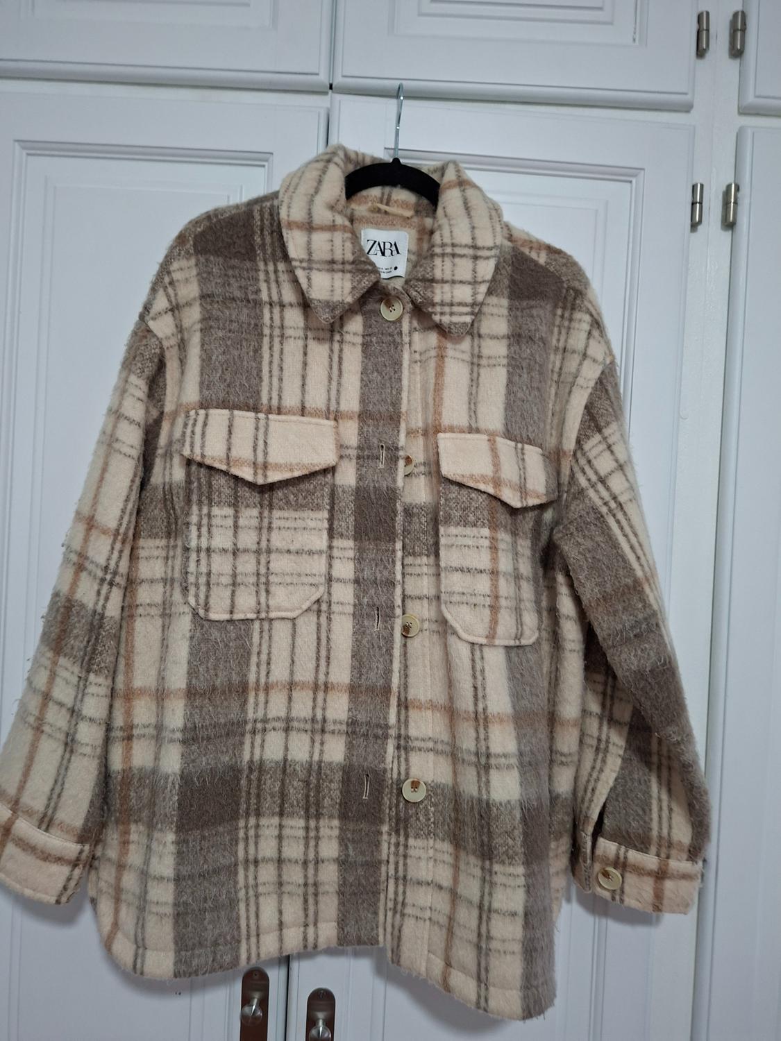 Brown and beige checked jacket hanging on a hanger in front of a white wardrobe. Visible label with the text "ZARA".