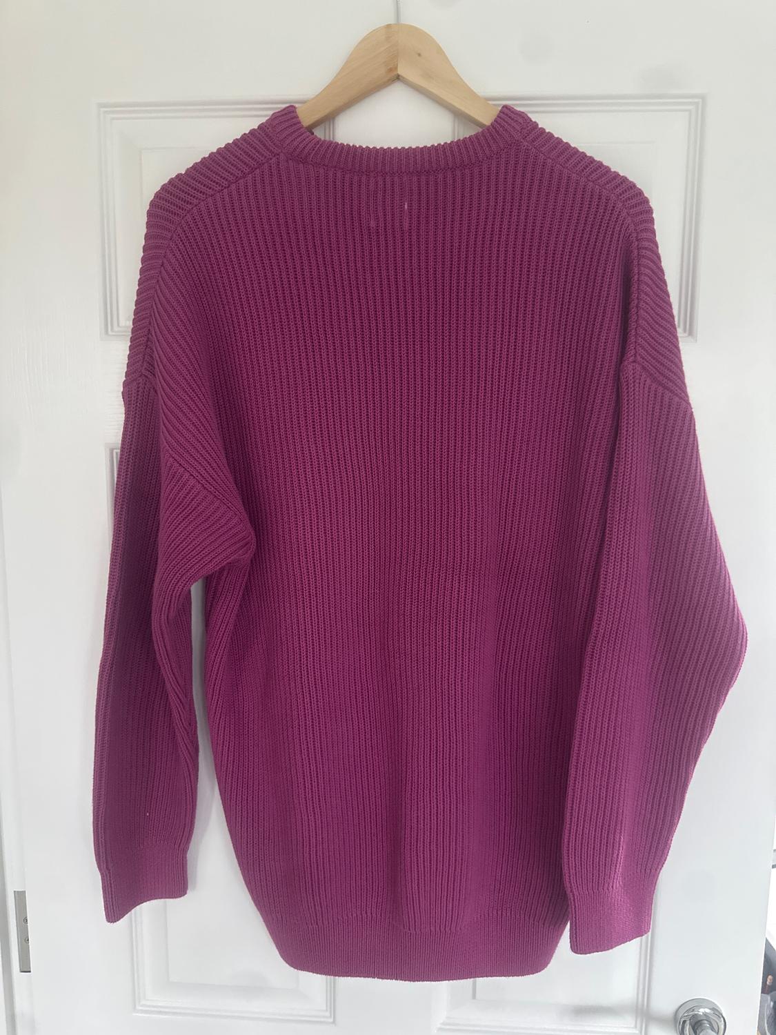 Fuchsia chunky knit jumper hanging on a wooden hanger over a white door.