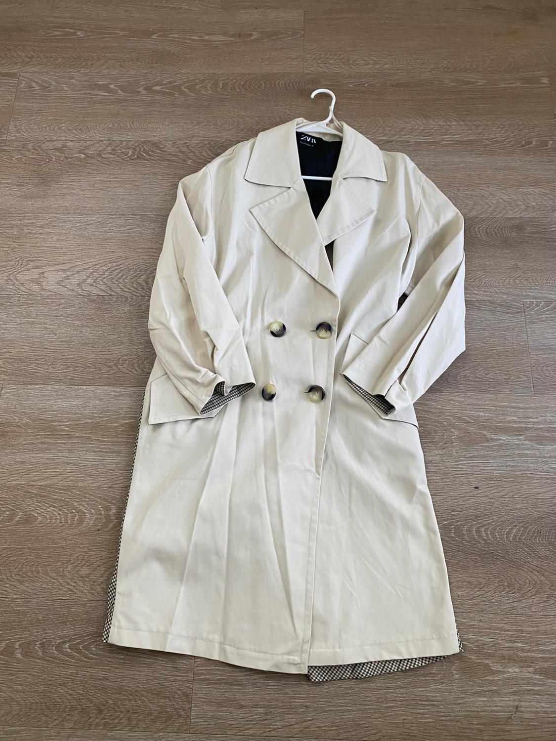 Double-breasted beige trench coat with wide lapels, hanging on a hanger over a wooden floor. Visible tag: "Zara".