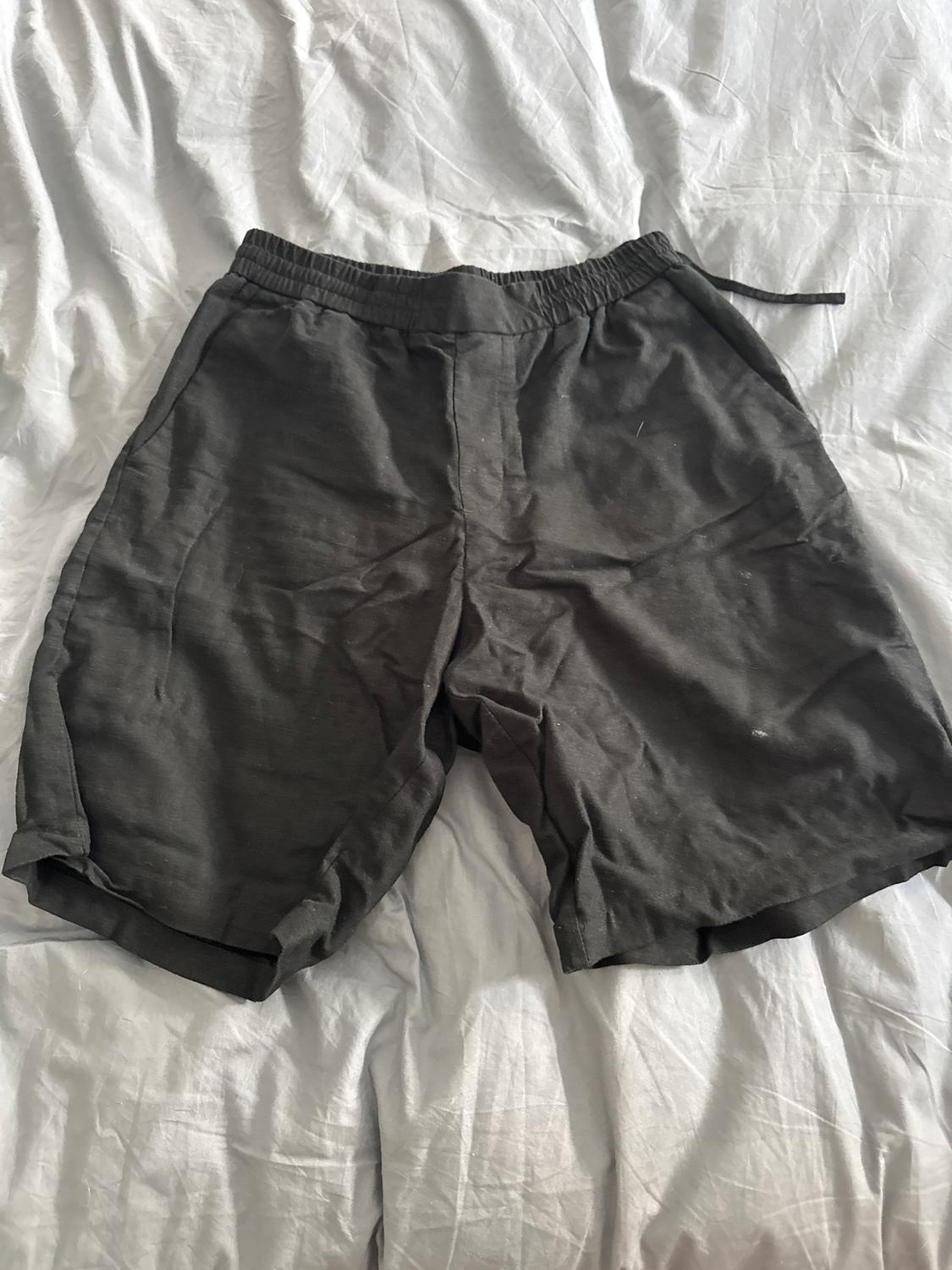 Dark shorts with an elasticated drawstring waist, placed on a bed with crumpled white sheets.