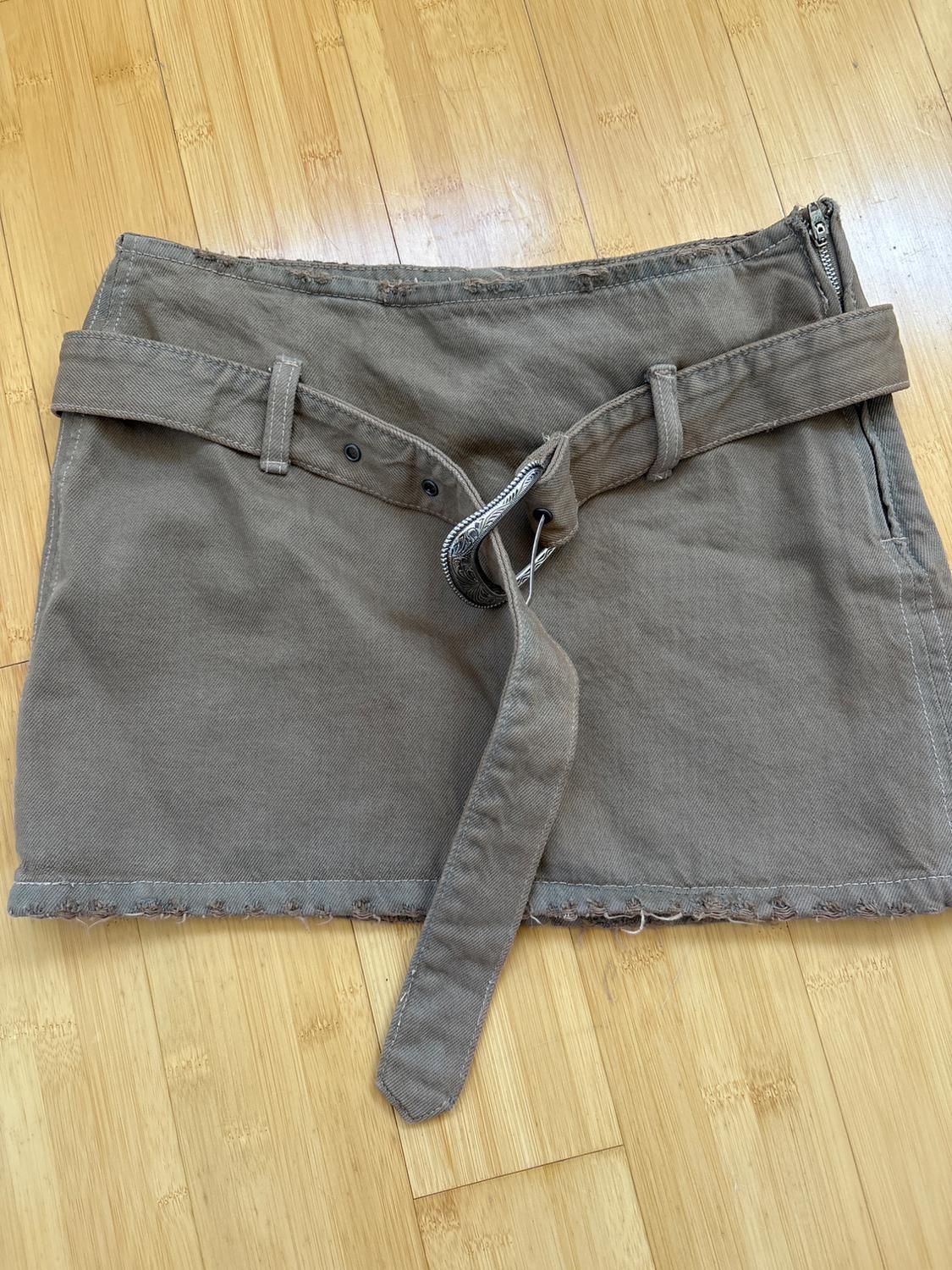 Brown mini skirt with a wide belt and metal buckle, side zipper closure, on a light wooden floor.