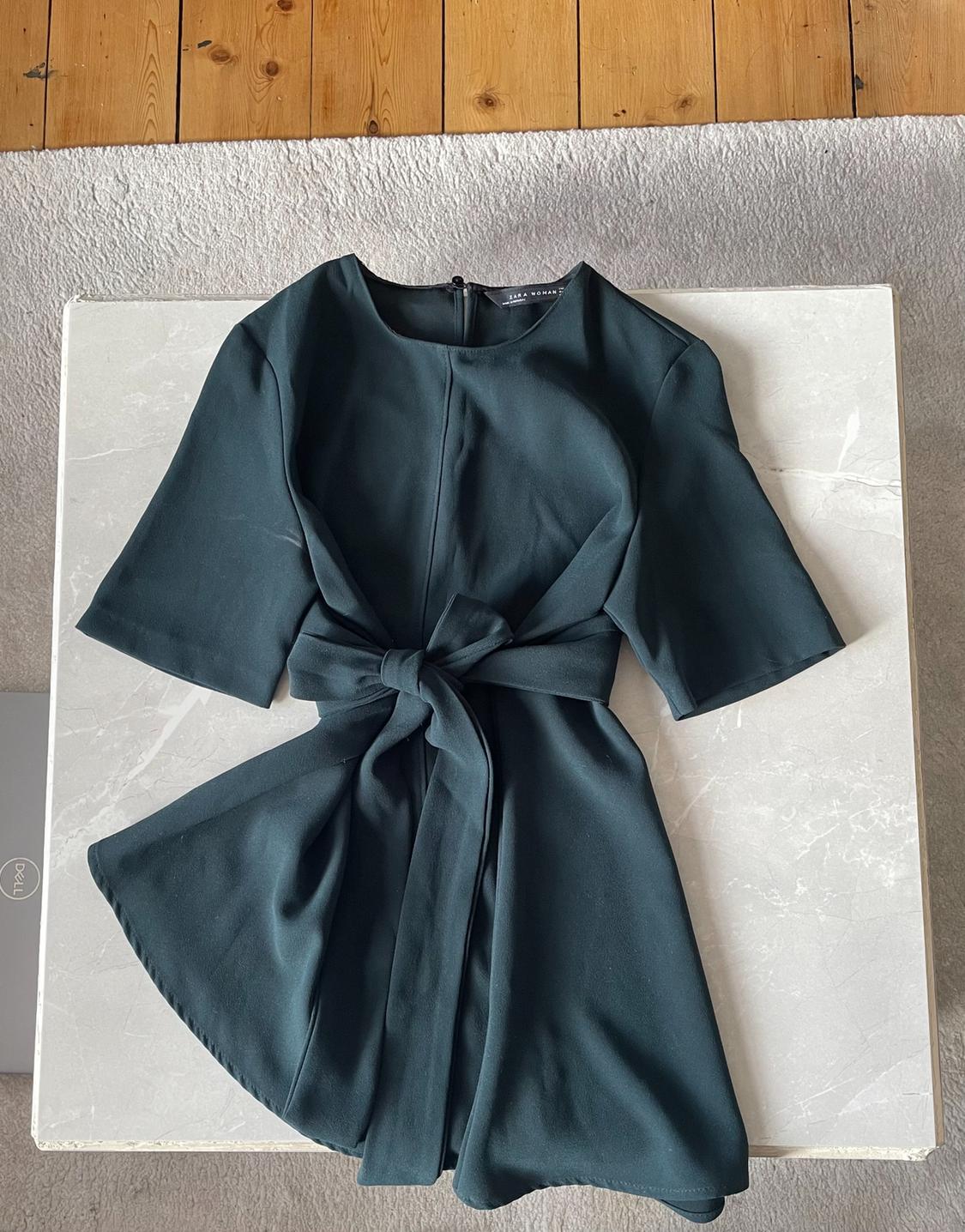 Dark green short-sleeved dress with a bow at the waist, on a light marble table. Visible text: ZARA WOMAN, DELL.
