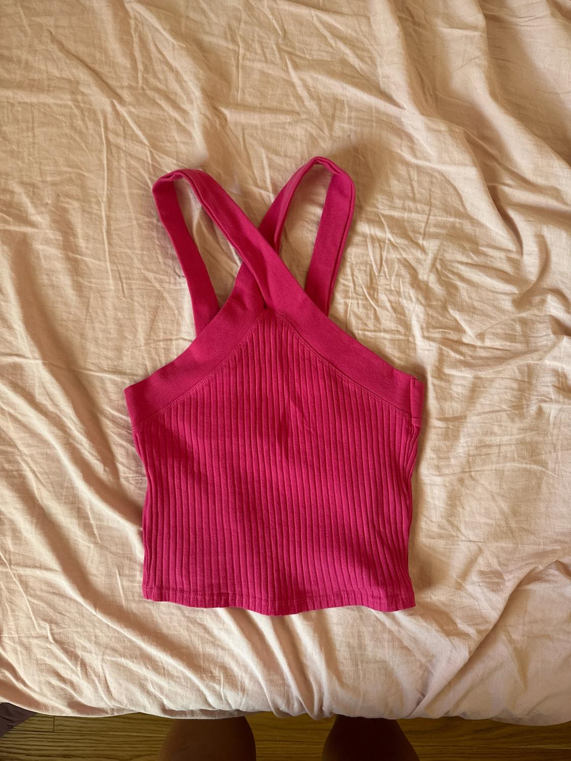 Fuchsia pink ribbed crop top with crossover straps on a crumpled beige sheet. A pair of legs can be seen at the bottom.
