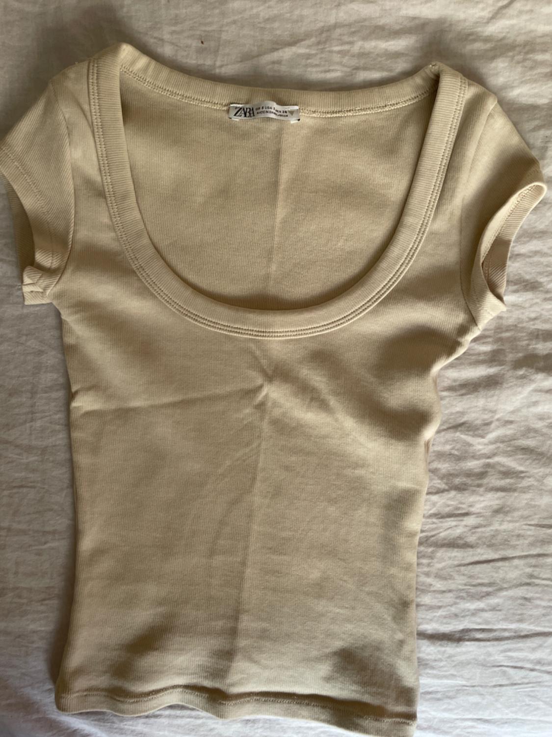 Beige short-sleeved T-shirt with a round neck on a white fabric surface. Visible label with text "ZYA".