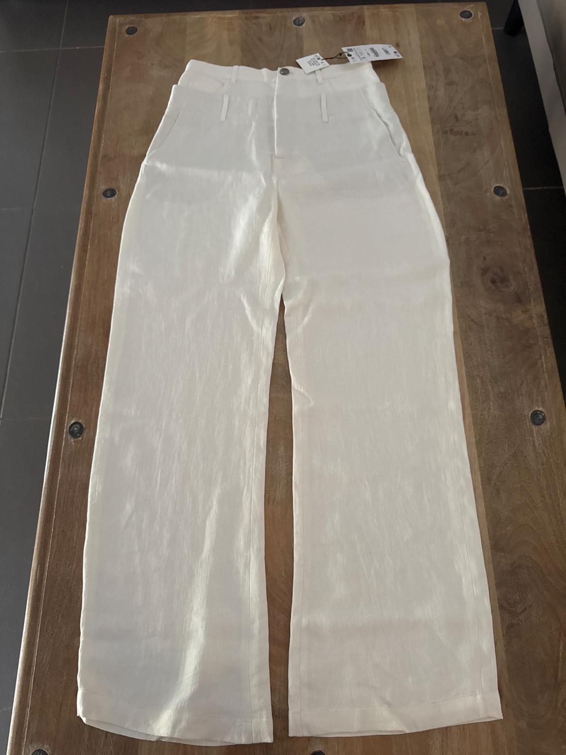 White, straight-leg trousers with front pockets, metal button and visible label, placed on a wooden table.