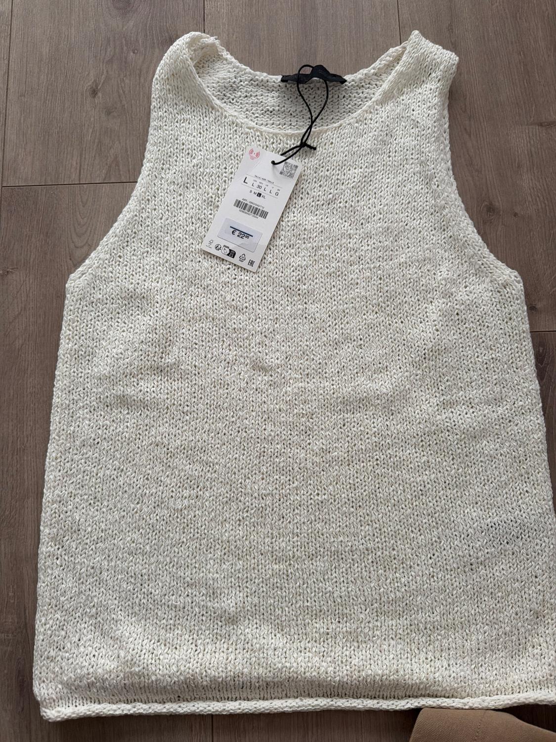 White knitted waistcoat on a wooden floor, with a price tag of 22.99 euros hanging from the neck.