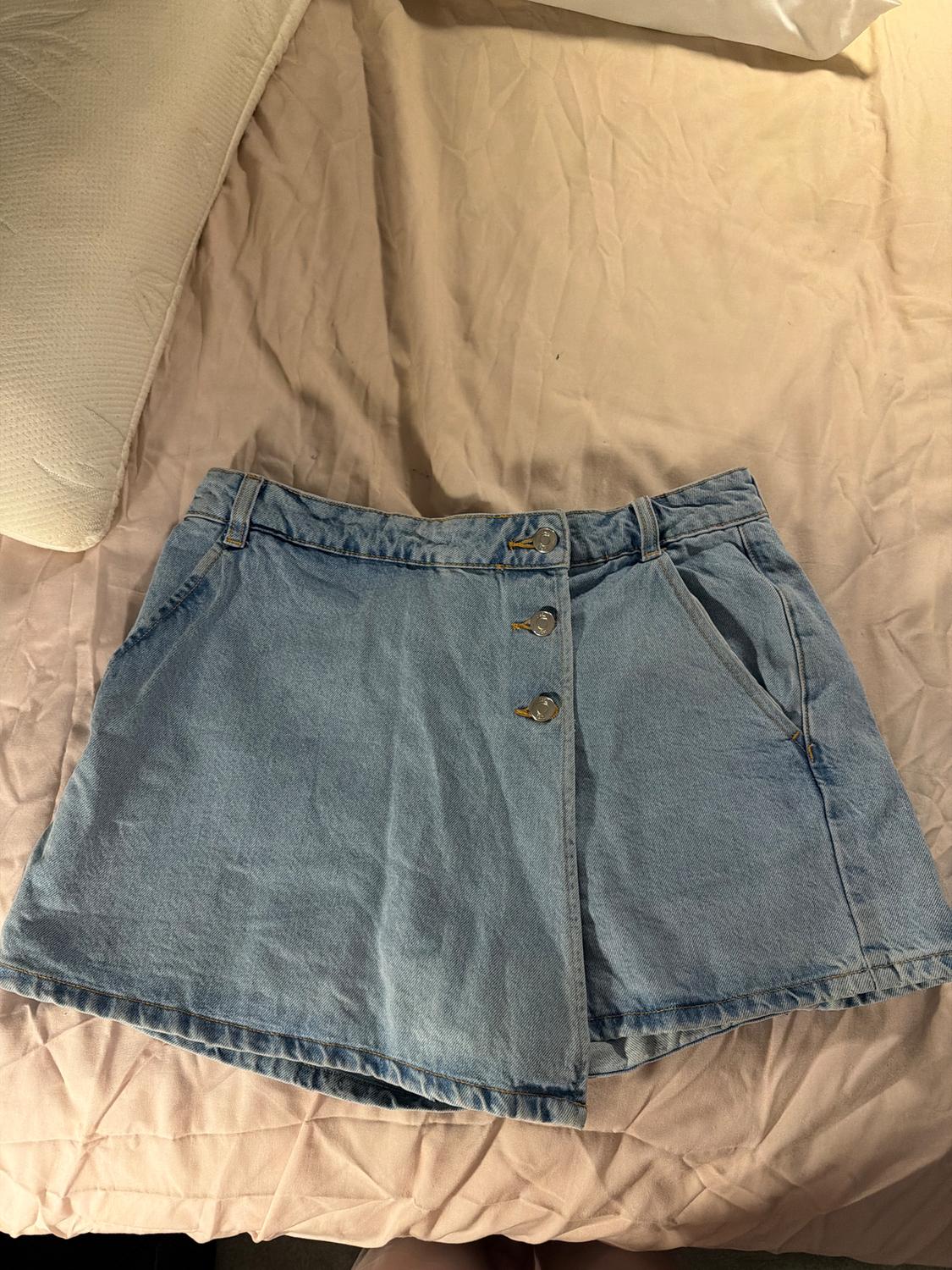 Light blue denim mini skirt with metal buttons, on a quilted beige bedspread with a white pillow beside it.