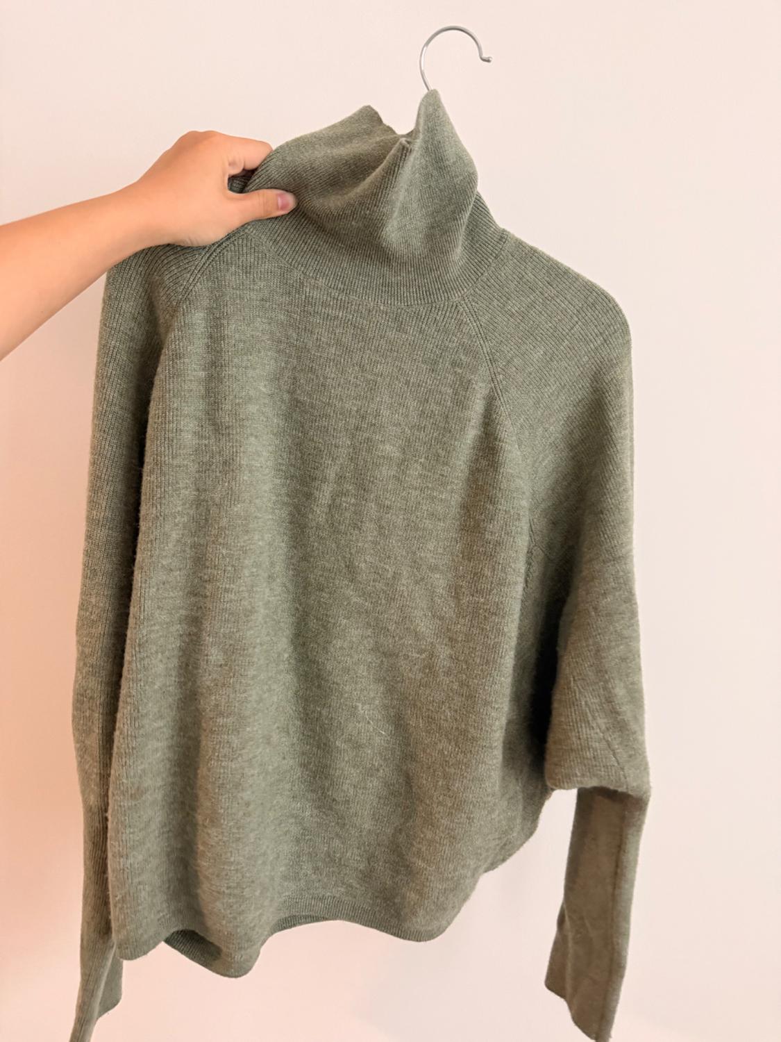 A hand holds an olive green knit turtleneck sweater on a hanger against a light background. Simple and relaxed composition.