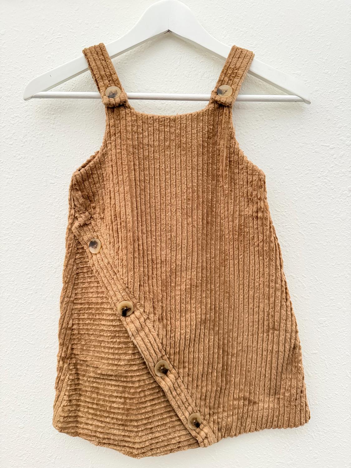 Brown corduroy dress with straps and buttons on the side, hanging on a white hanger against a white background.