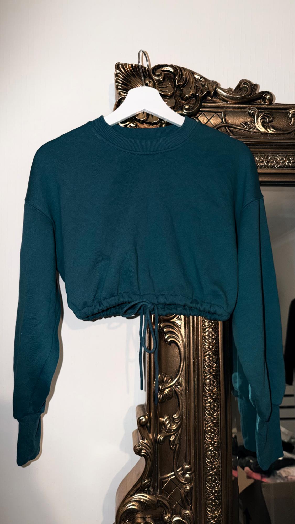Petrol Blue Cropped Sweatshirt hanging on a white hanger in front of an ornate gold-framed mirror.