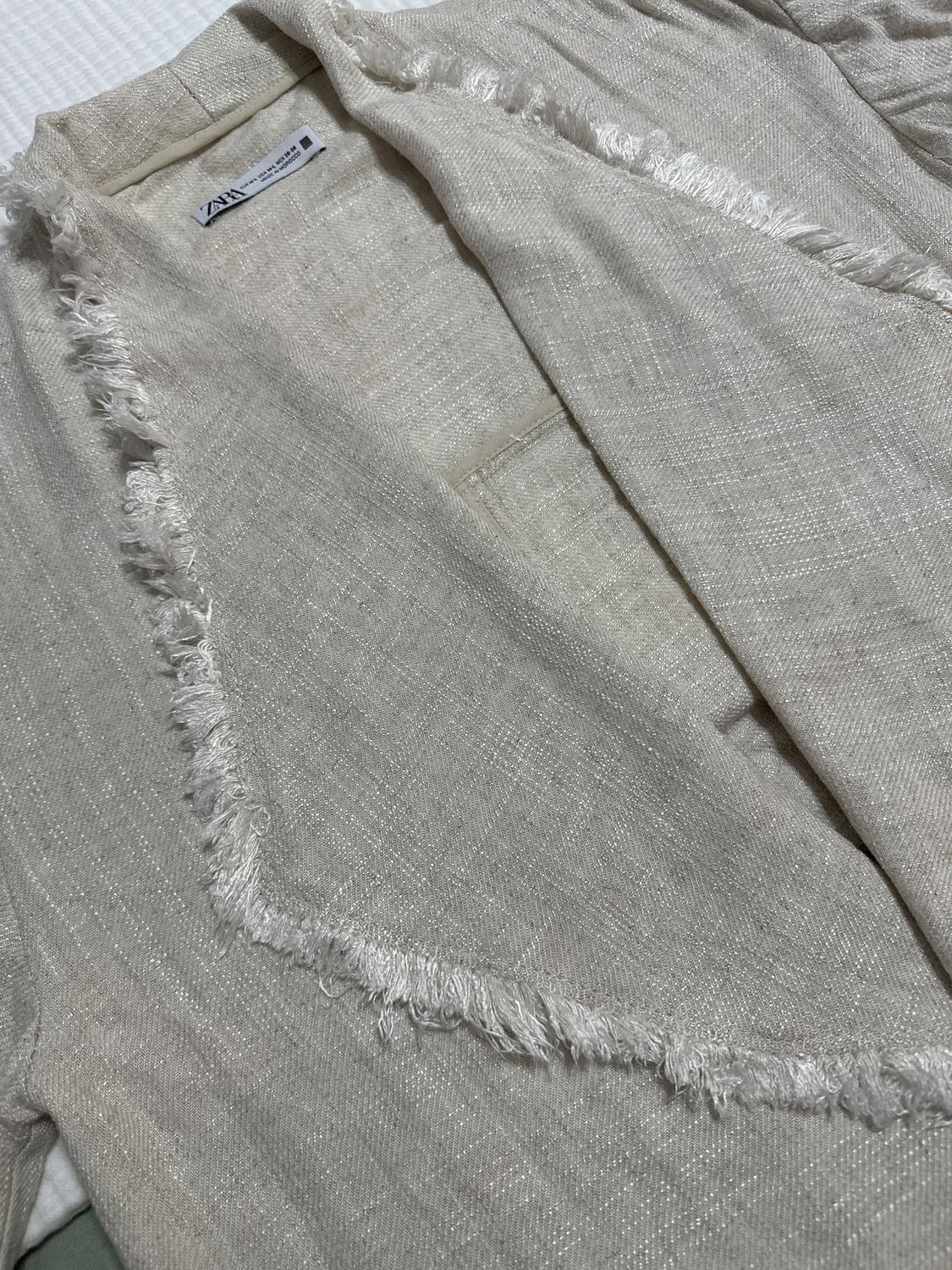 Beige jacket with texture and frayed edges. Visible tag with text: "ZARA". White background.