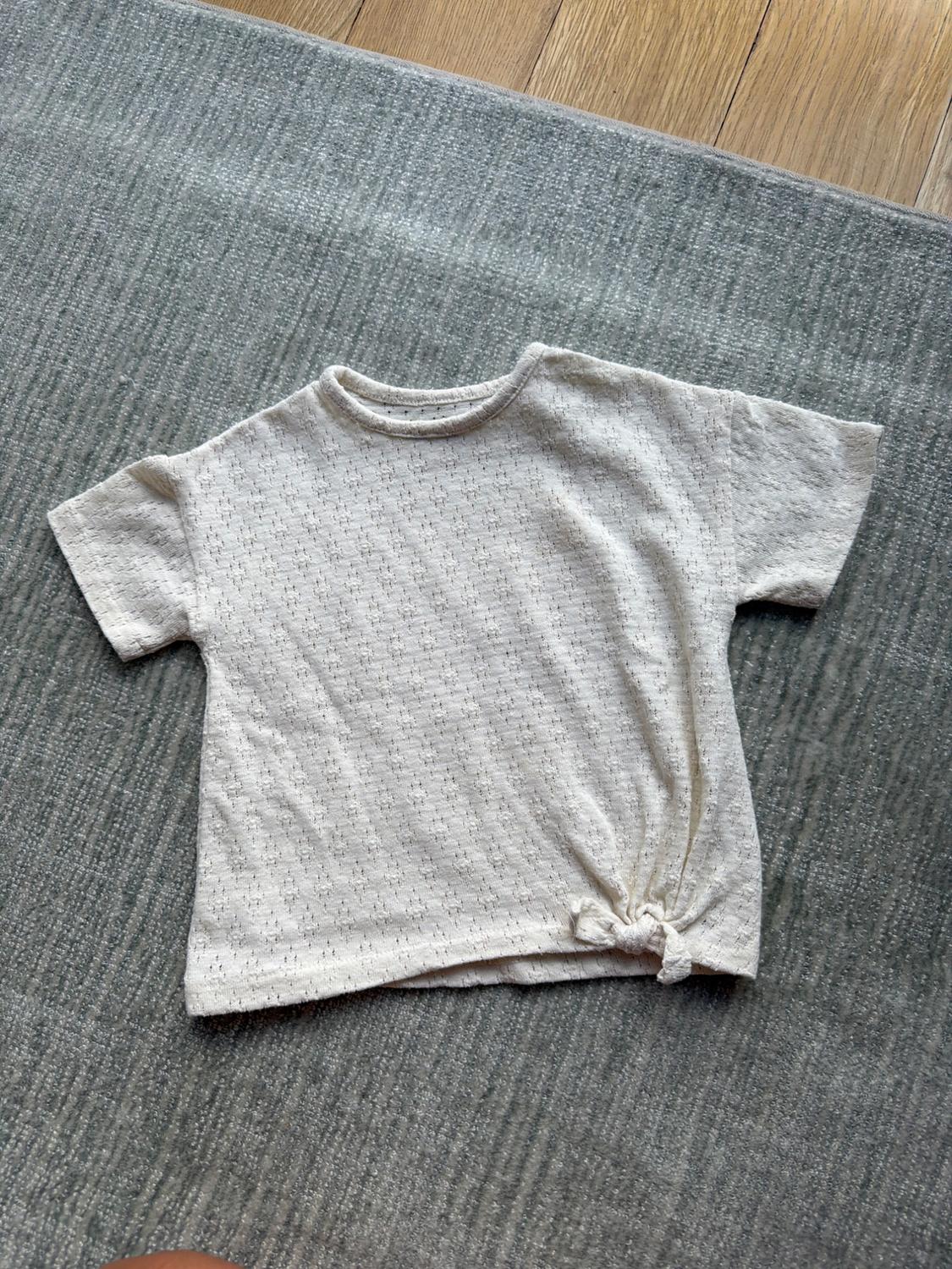 Beige, short-sleeved, textured T-shirt, tied at the side, on a grey rug and light wood floor.