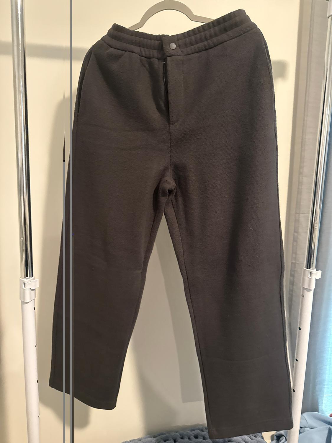 Brown trousers hanging on a hanger, with a button at the waist, against a light background and blue curtains to the right.