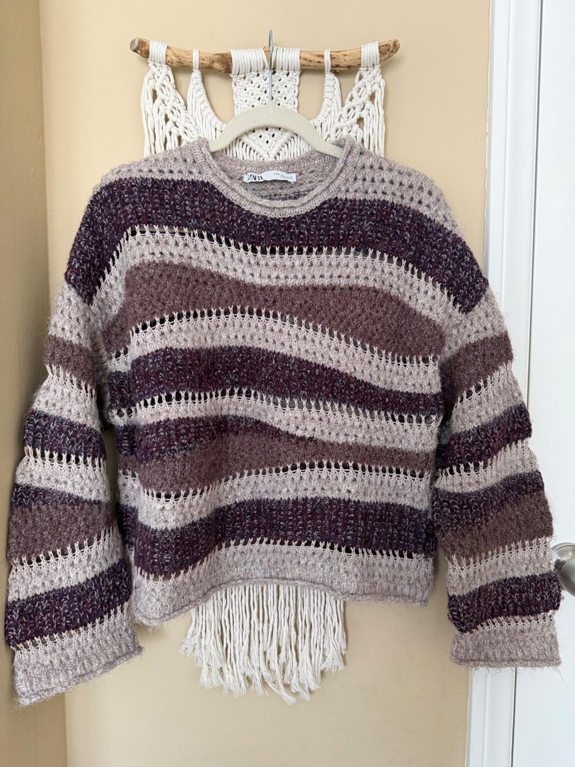 Knit sweater with horizontal stripes in brown and beige tones, hanging on a hanger against a white macrame background.