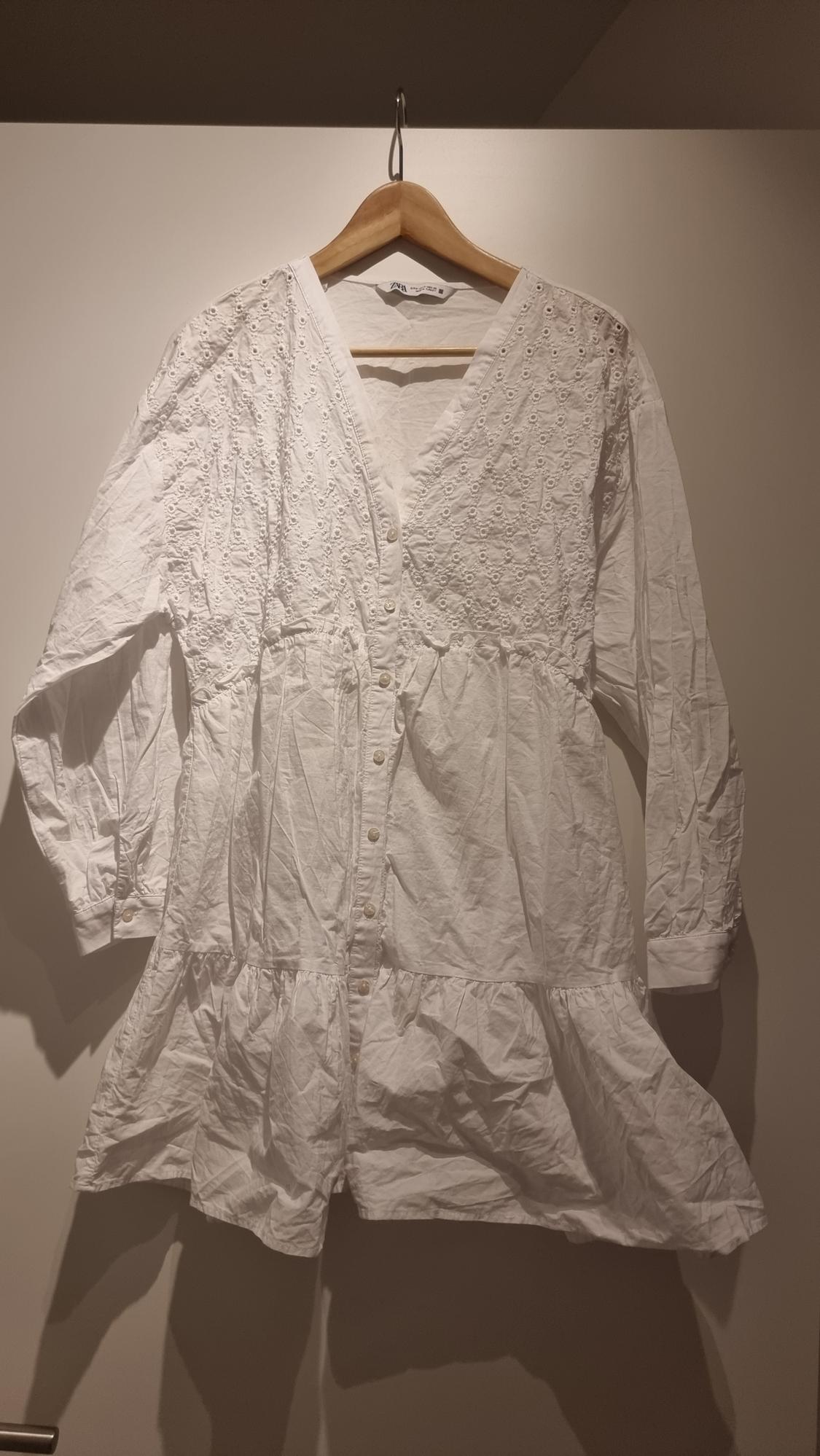 White cotton dress with floral embroidery, long sleeves and front buttons, hanging on a wooden hanger against a neutral background.
