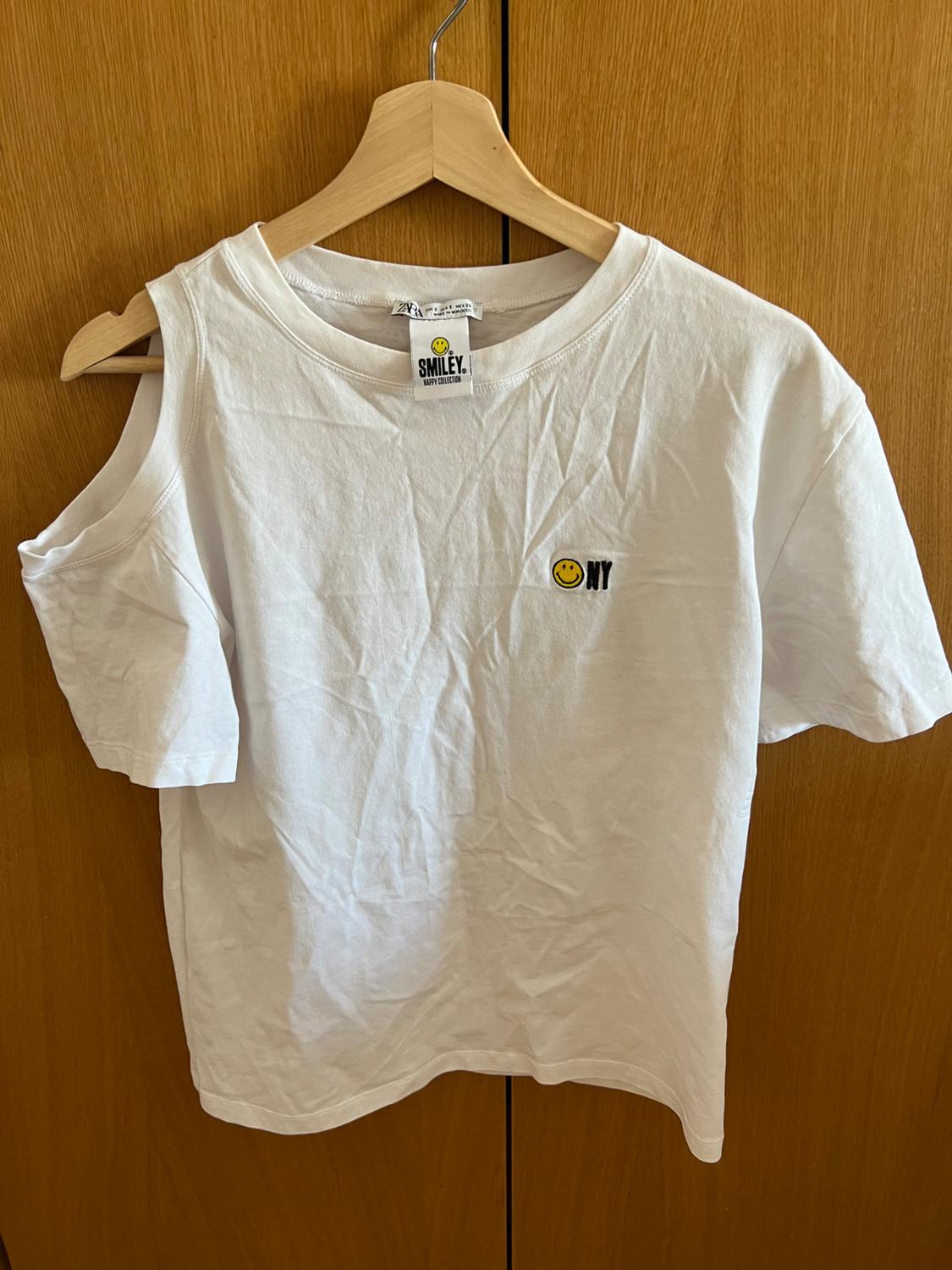 Casual white T-shirt on a wooden hanger with one shoulder off. Text: ZARA, SMILEY HAPPY COLLECTION, NY and smiley face logo.