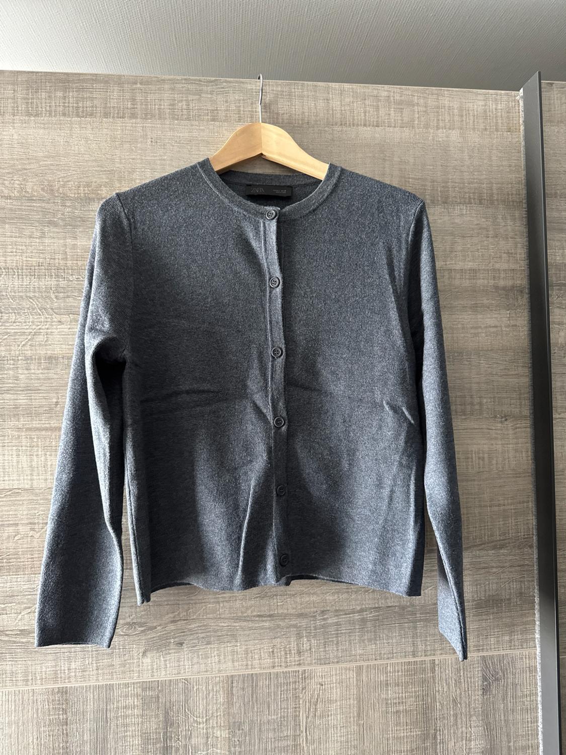 Grey long-sleeved button-up cardigan, hanging on a wooden hanger against a wooden background. ZARA label.