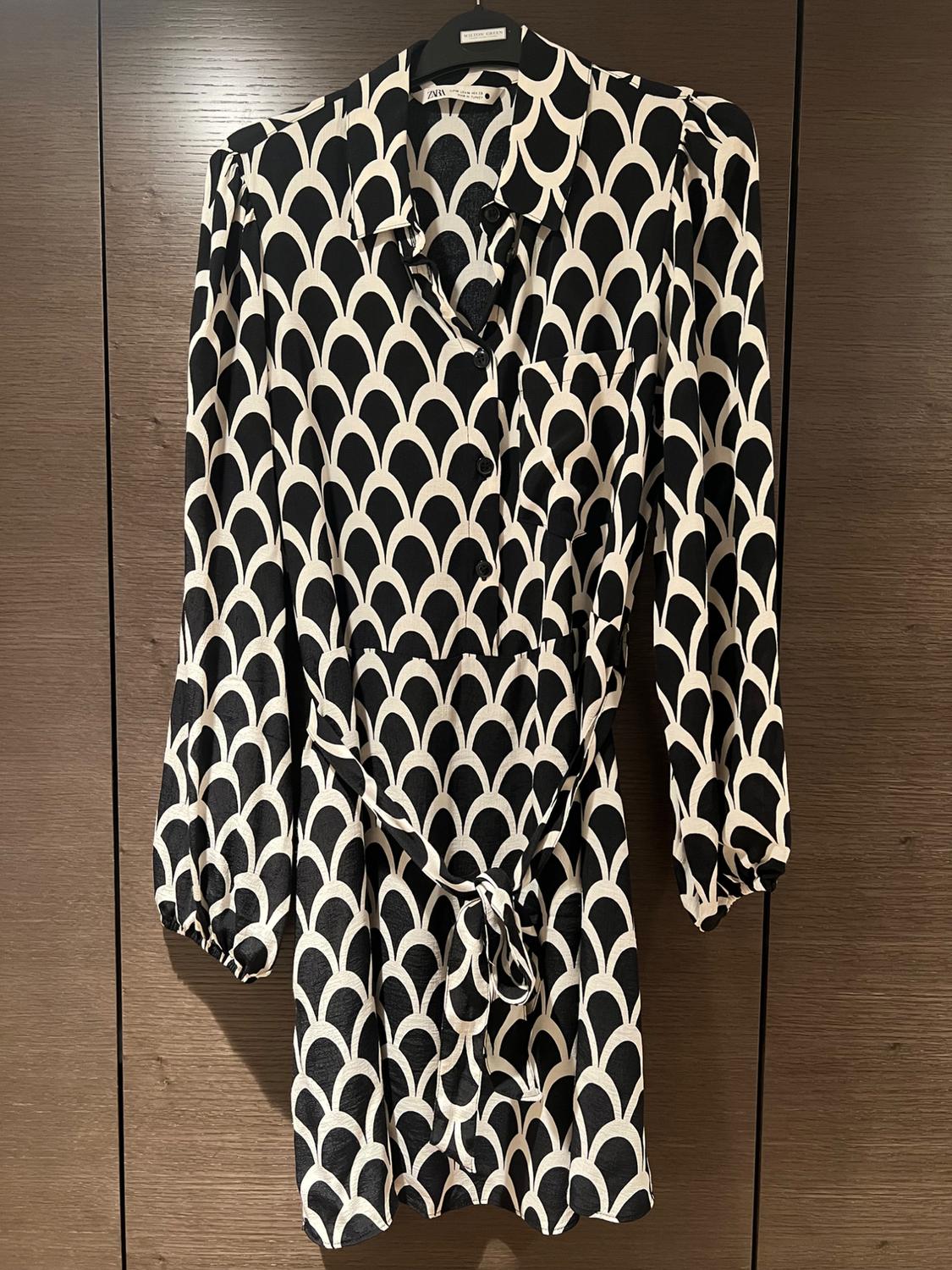 Blouse hanging with black and white geometric print, dark wood background. Visible label: "Zara".