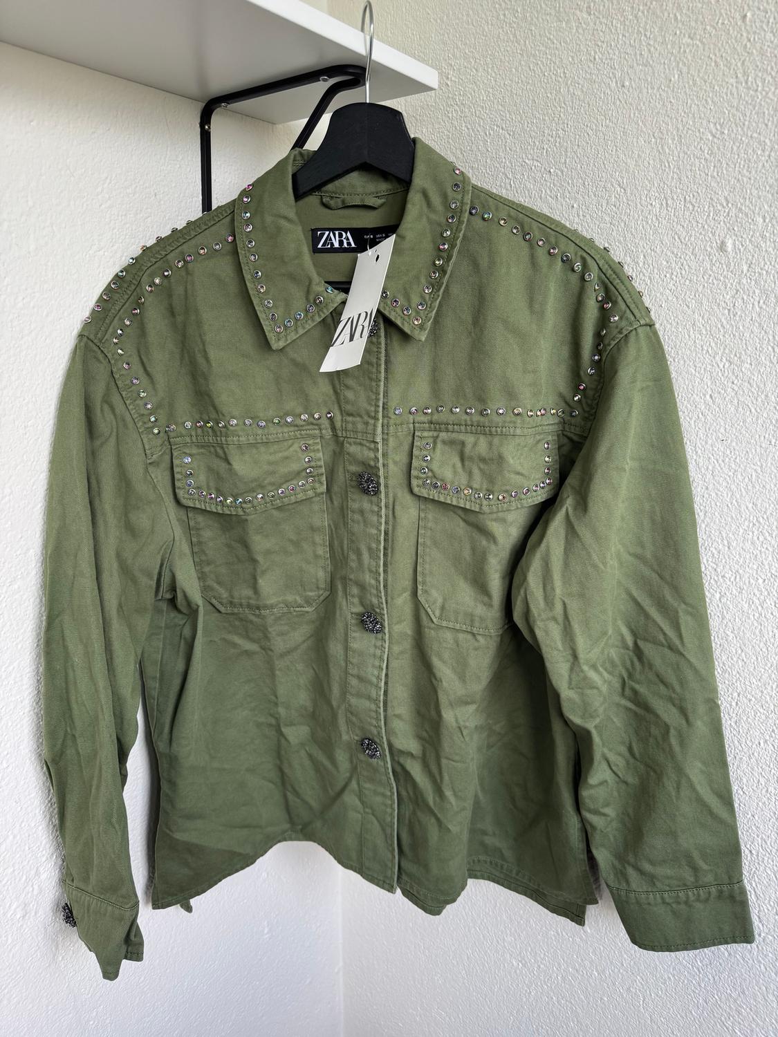 Olive green jacket with studs on shoulders and pockets, hanging on a black hanger with a Zara tag, on a white background.