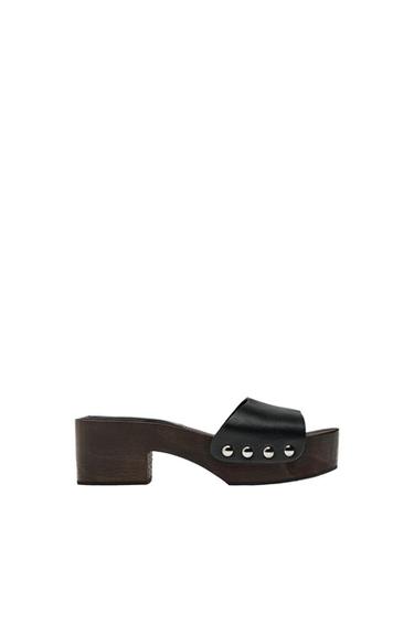 Low-heeled sandal with a dark wood sole and a wide black strap with metallic studs, on a white background.