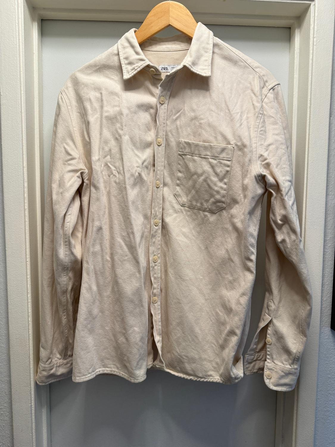 Long-sleeved beige shirt hanging on a wooden hanger. It has a chest pocket and front buttons. Visible tag: Zara.