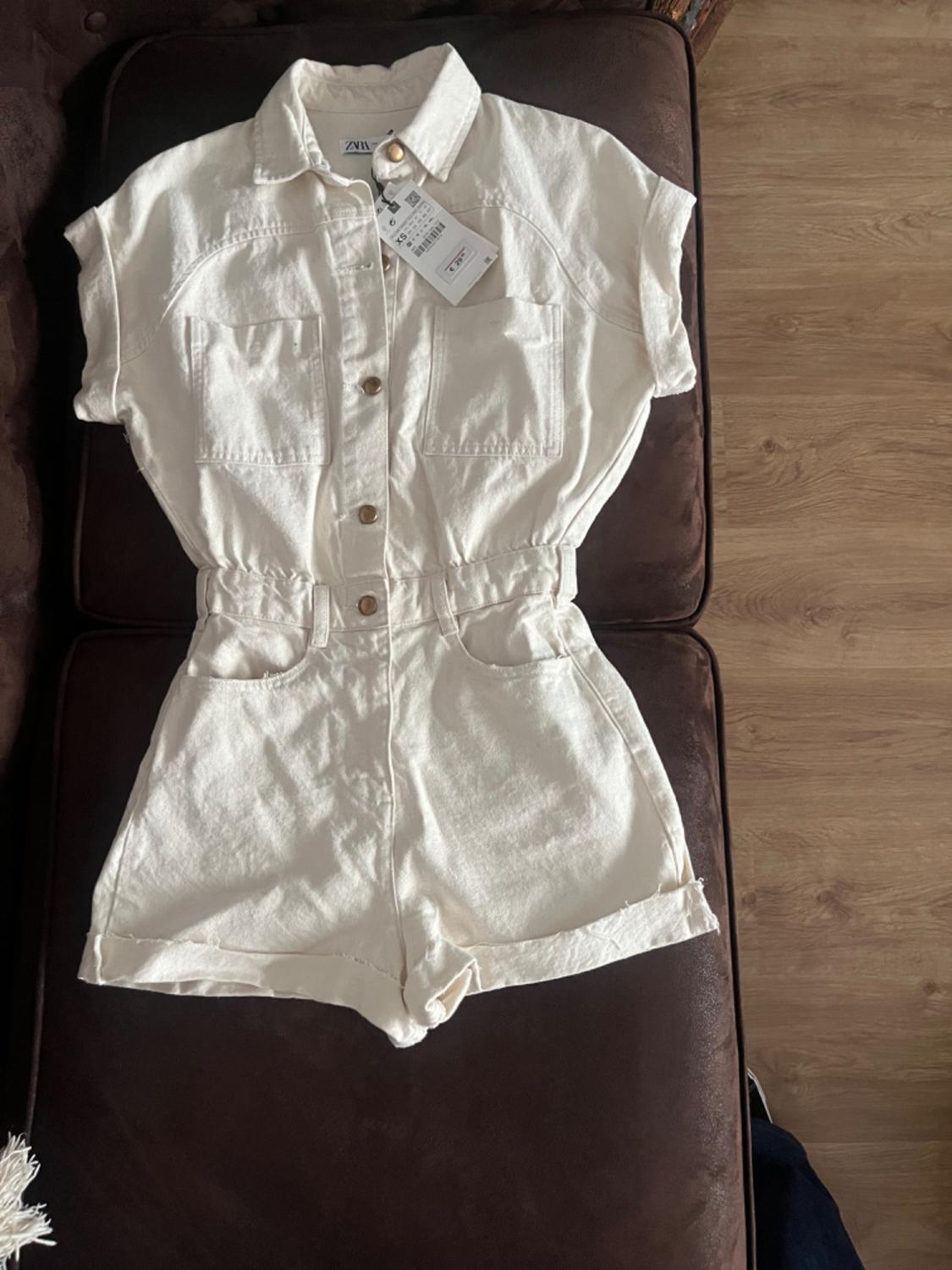 White playsuit with buttons and front pockets, placed on a brown sofa. Visible label with text.