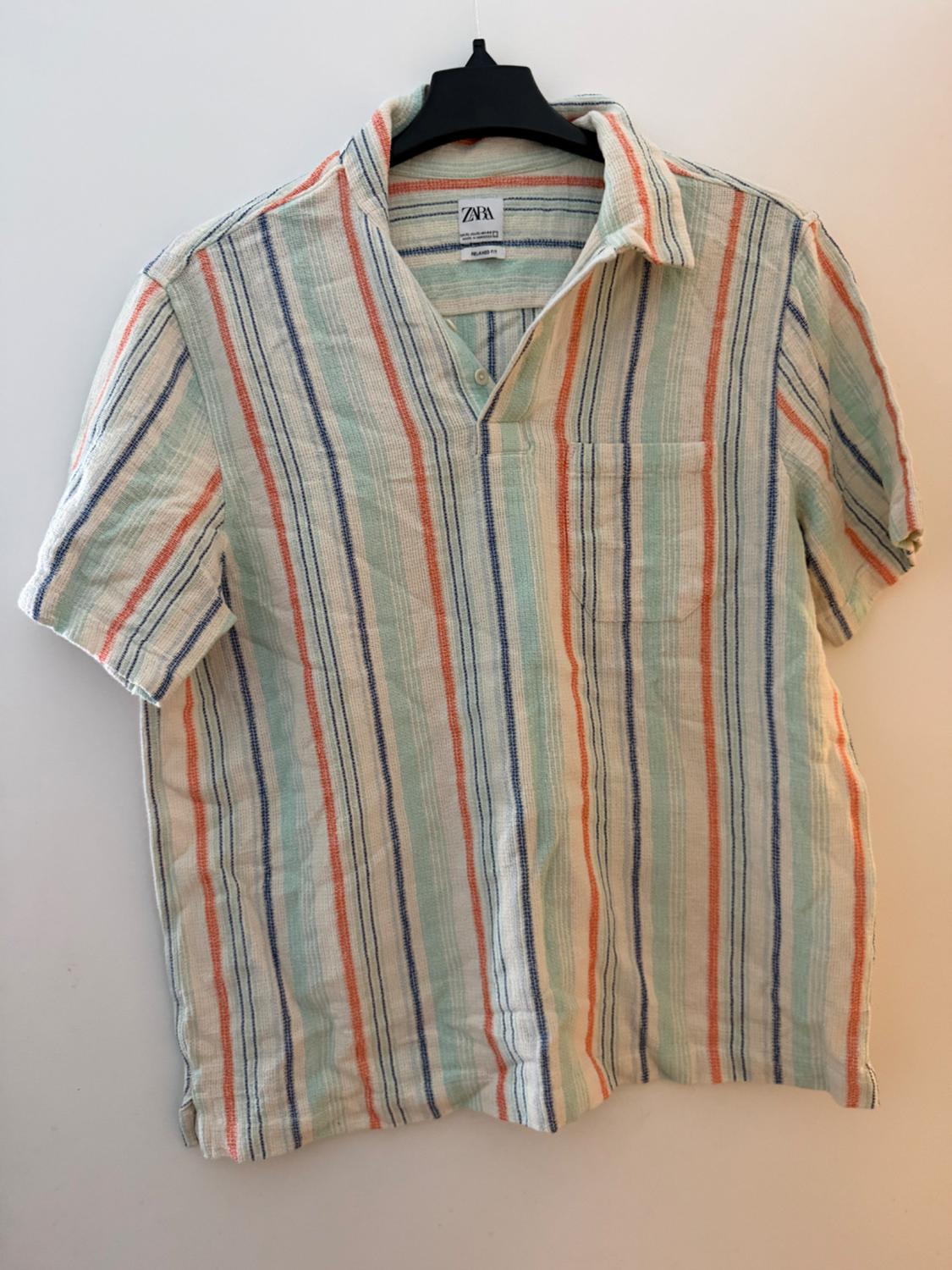 Short-sleeved shirt with a pocket and orange, light blue and blue vertical stripes on a black hanger. Label with text ZARA.