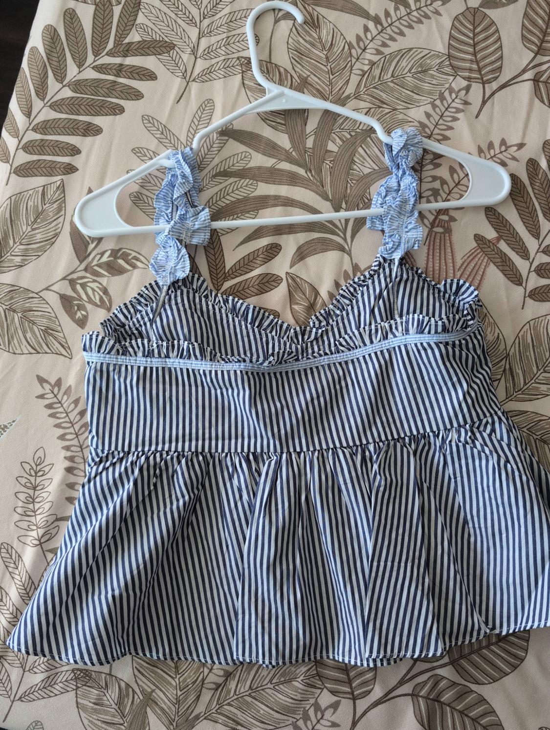 Blue and white striped top with ruffle straps on a white hanger, over a beige leaf-print fabric.