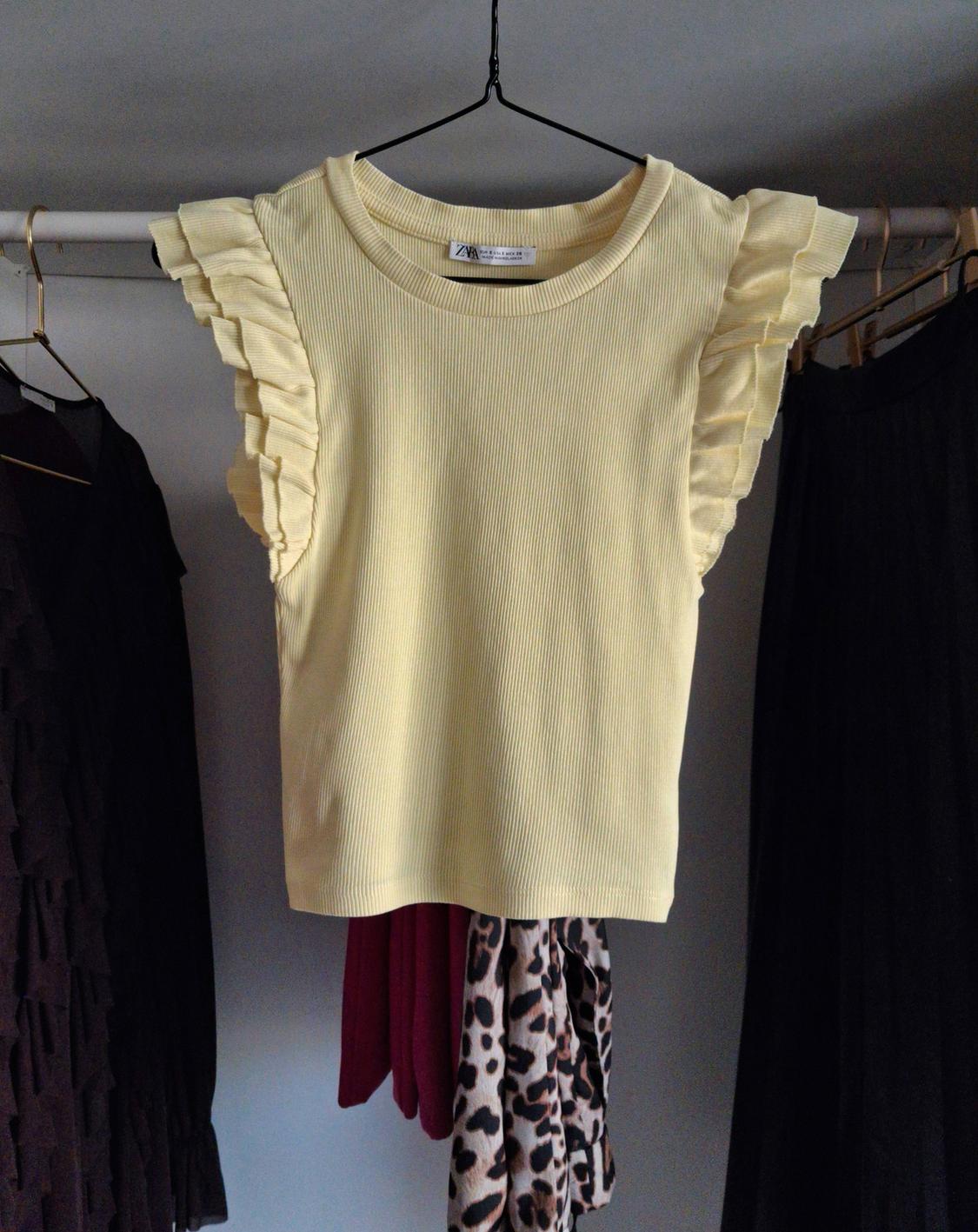 Yellow blouse with ruffled sleeves hanging on a clothes rail, surrounded by dark garments and animal print.