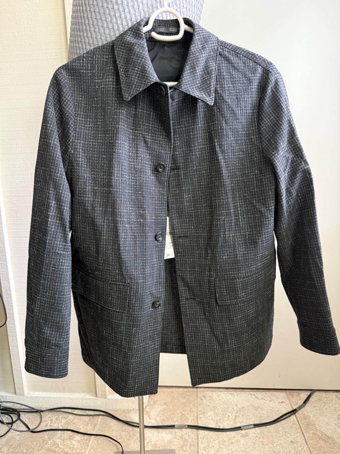 Dark grey jacket with a white grid pattern, hanging on a hanger on a metal stand. ZARA label visible inside.