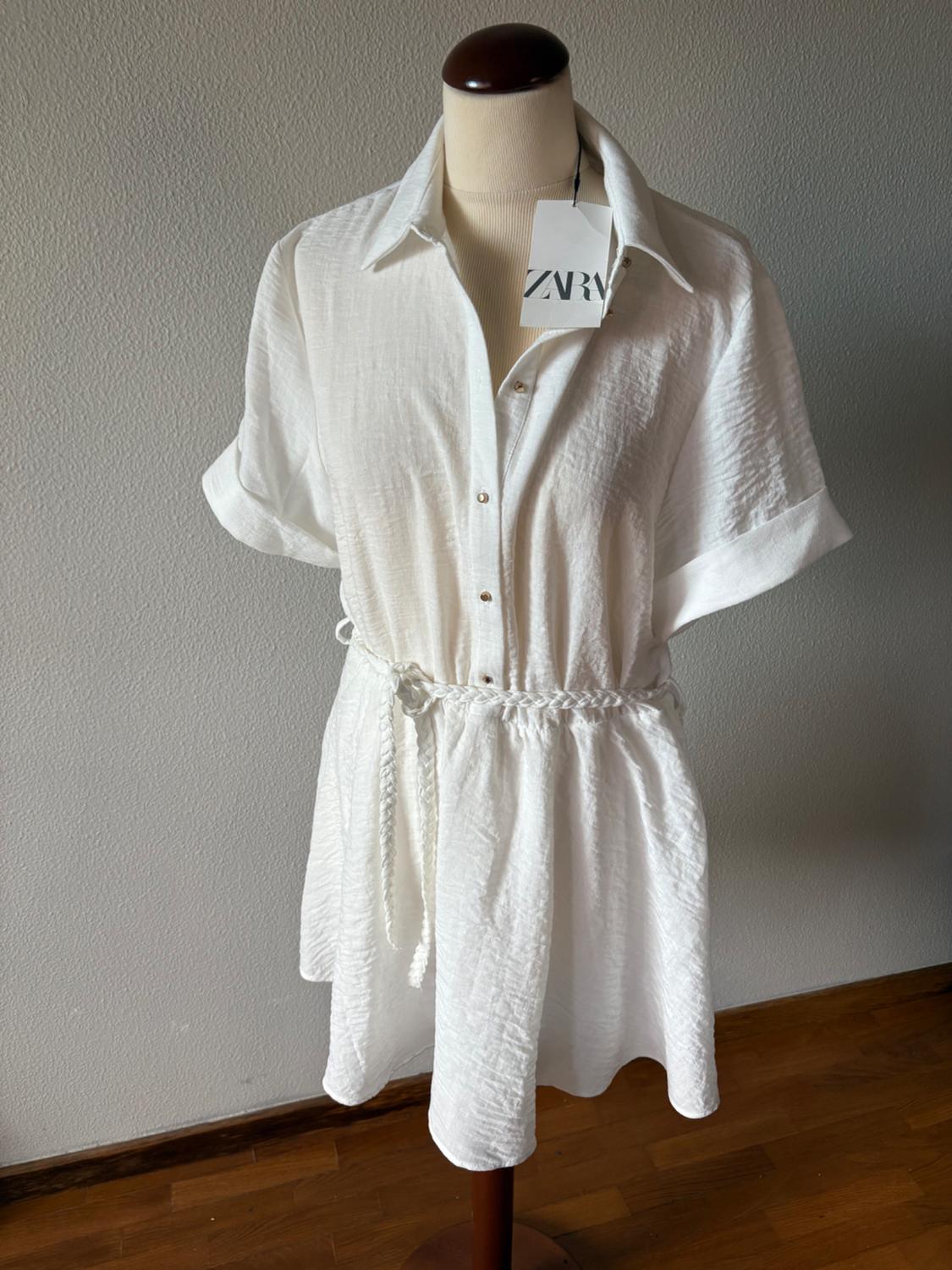 Mannequin wearing a textured white shirt dress, short sleeves and a braided belt. Label with the text ZARA. Grey wall and wooden floor.