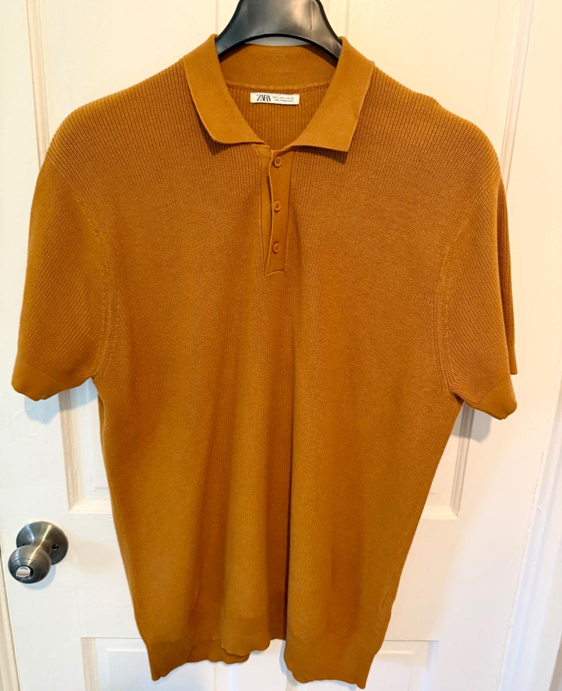 Mustard-coloured polo shirt hanging on a black coat hanger over a white door. Label visible with the brand "Zara".