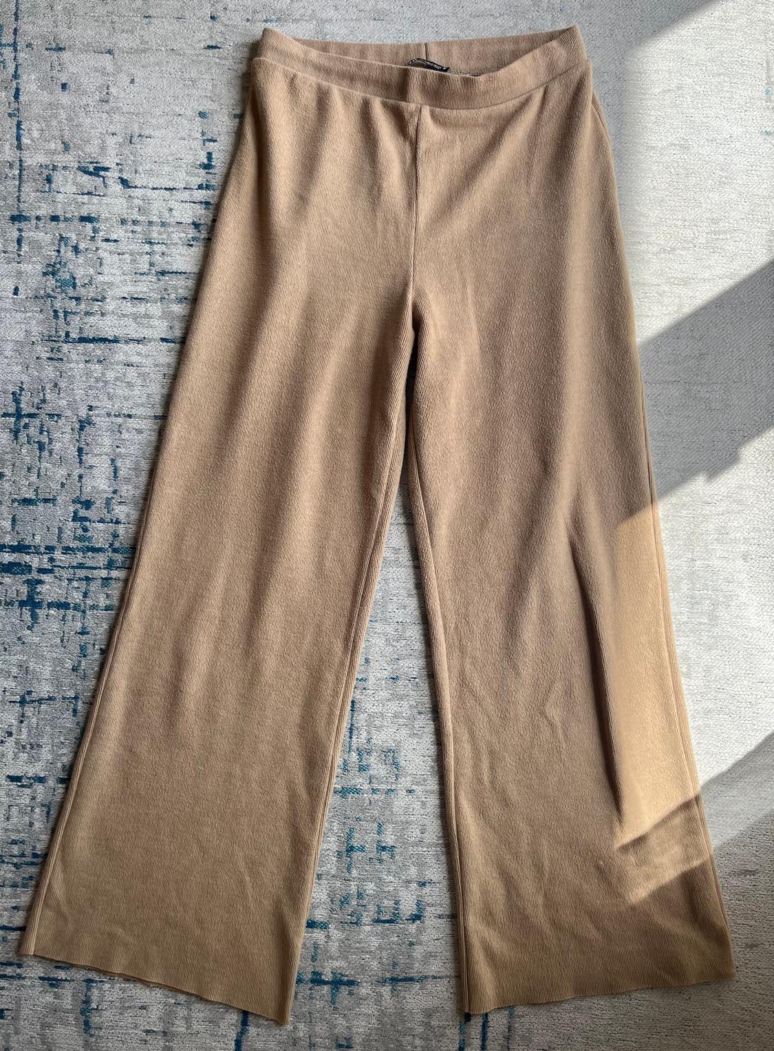 Beige wide-leg trousers on a grey rug with blue accents. Sunlight creates shadows on the fabric.