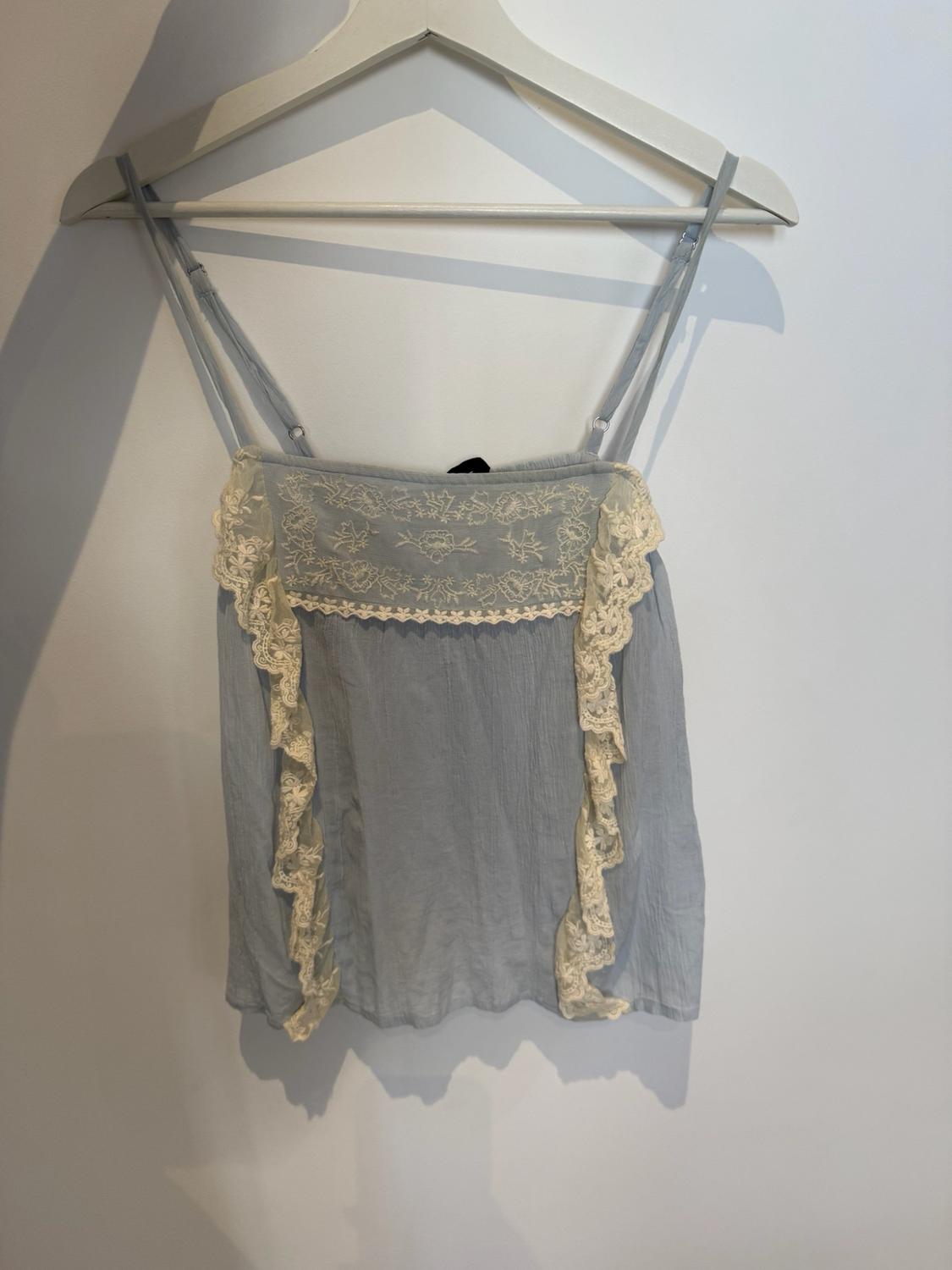 Light blue camisole top with thin straps on a white hanger. Its design stands out with cream embroidered lace at the neckline and frills on the sides.