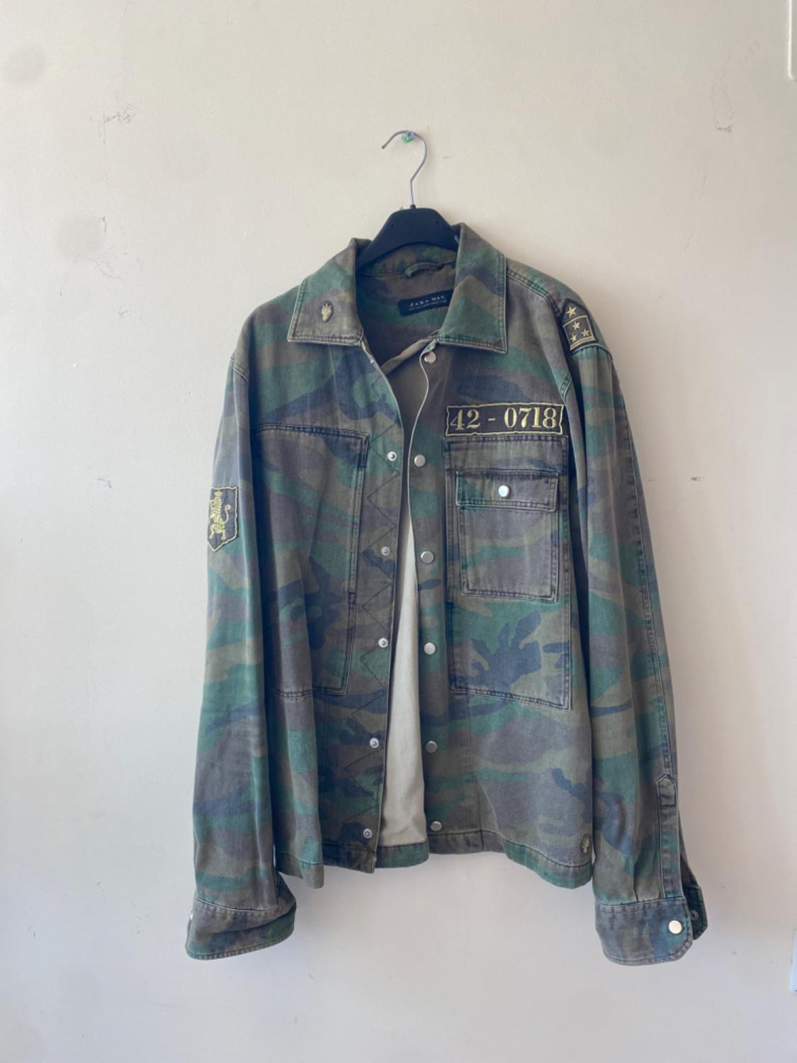 Camouflage jacket hanging on a black hanger, with patches and text "42-0718" on the chest, on a white background.
