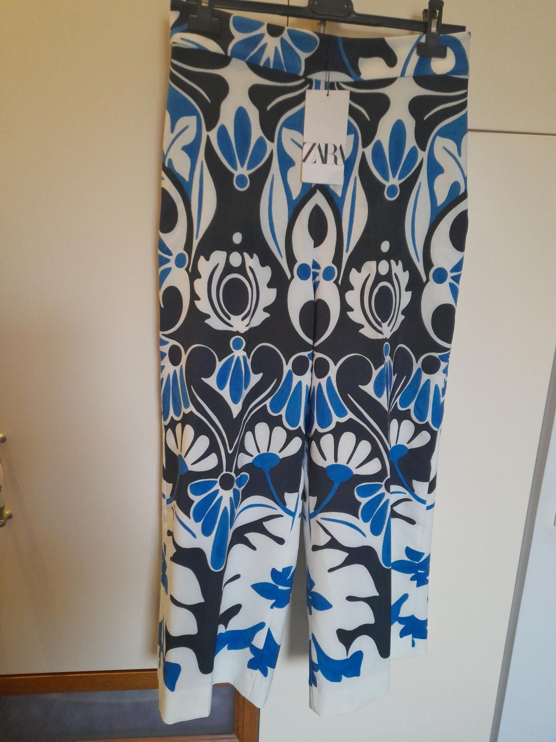 Trousers hanging with a floral print in blue, black and white. Visible label with the text "ZARA". Light-coloured wall background.