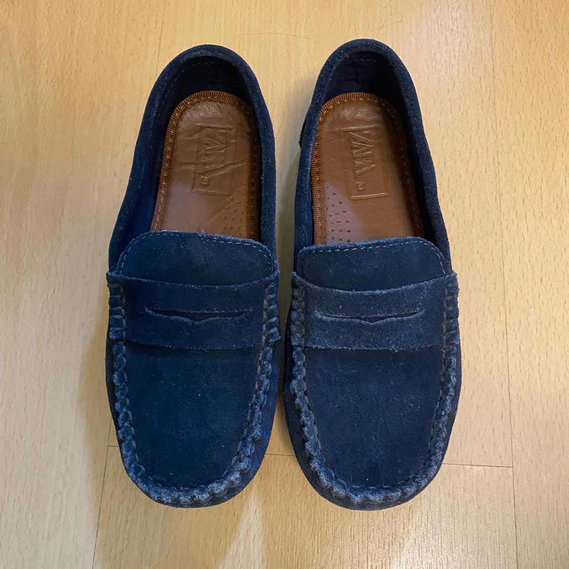Navy suede loafers with visible stitching, brown interior with embossed logo, on a light wood background.