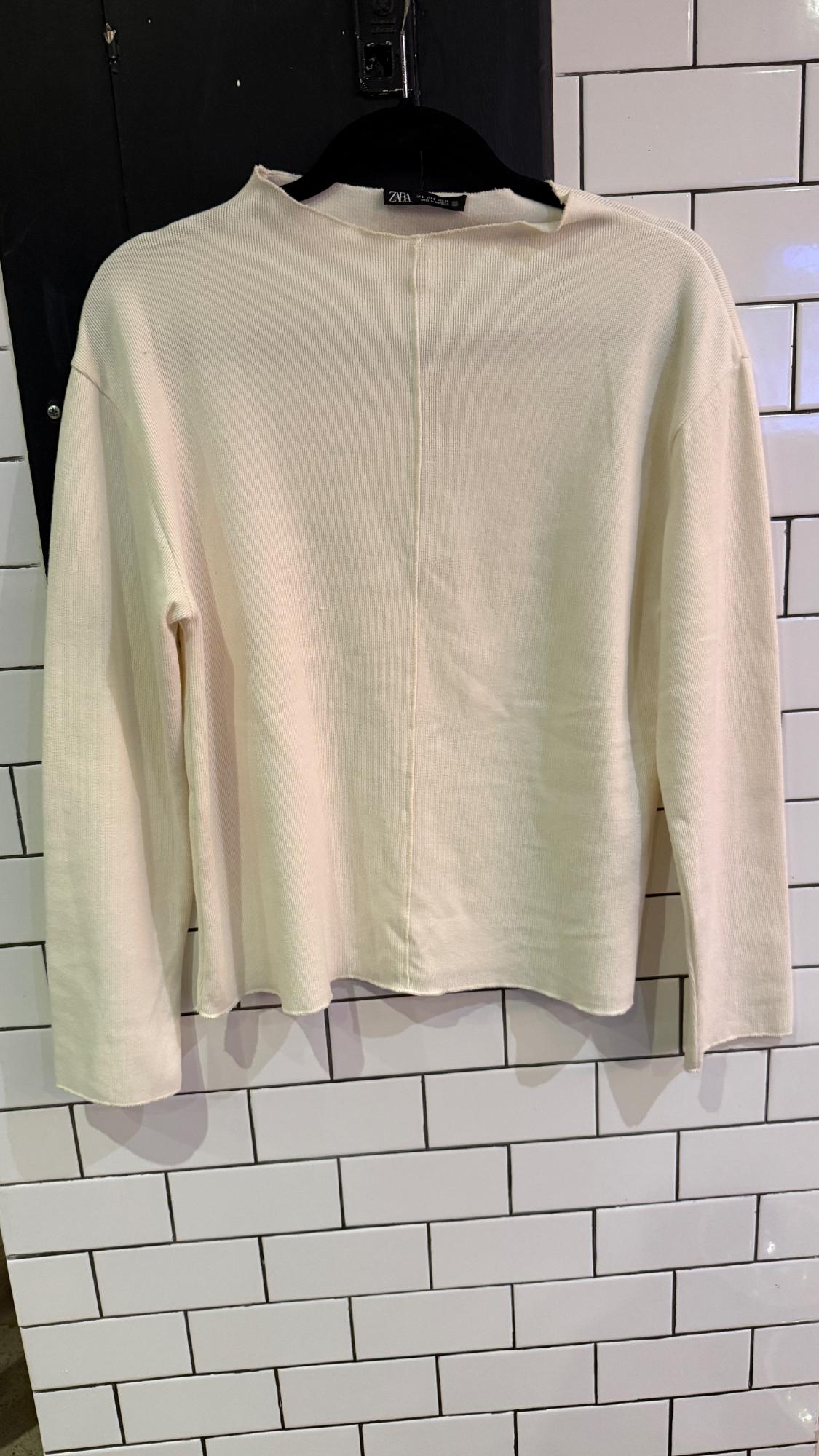 Beige long-sleeved sweater hanging on a black hanger in front of a white tiled wall. Visible tag: "Zara".