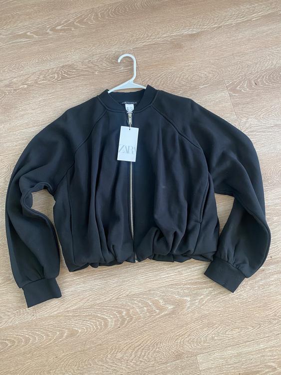 Black jacket with zipper and hangtag on a white hanger, placed on a light wood floor.
