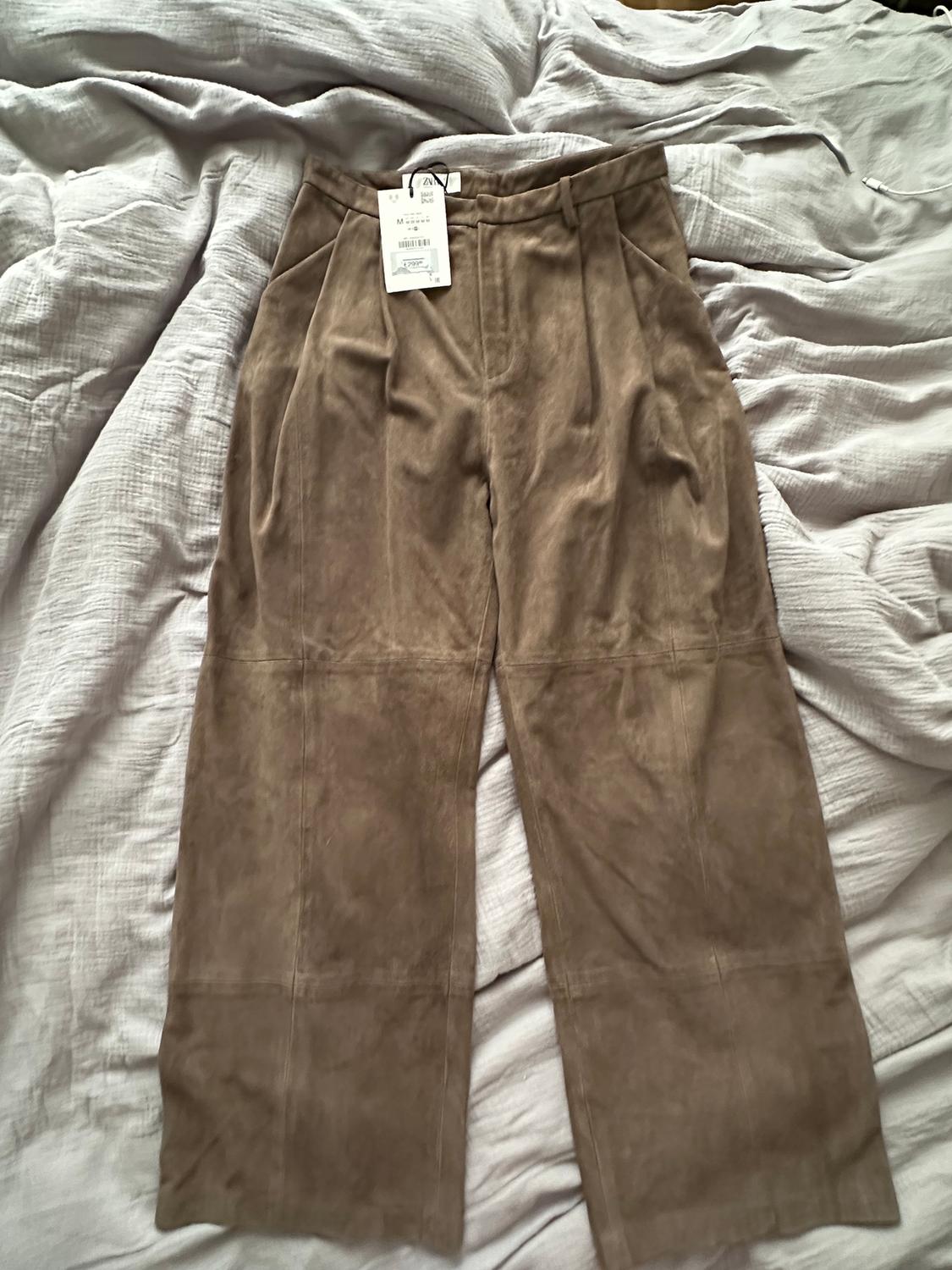 Brown, soft fabric trousers on a bed with wrinkled sheets. Label visible on the waistband.