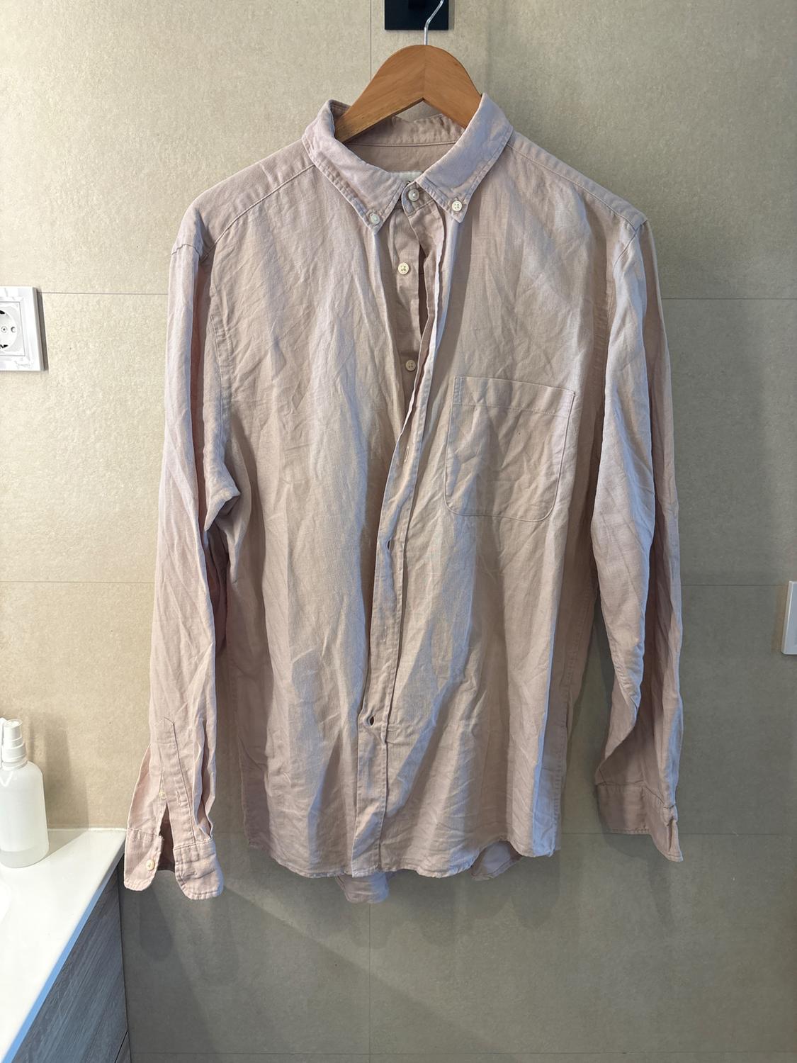 Light pink long-sleeved button-up shirt with a crinkled appearance, hanging on a wooden hanger against a beige tiled wall.