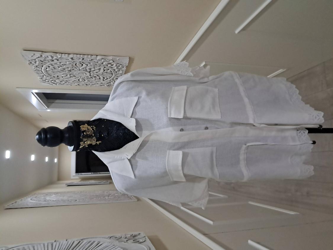 Mannequin in a white linen jacket with large pockets and lace details on the sleeves, against a decorated beige wall background.