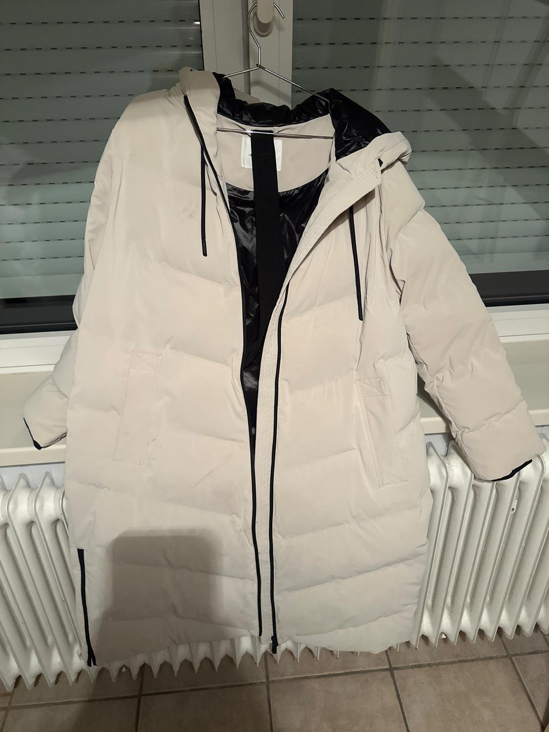 Beige long padded coat with a hood and black lining, hanging on a hanger in front of a window with blinds and a white radiator.
