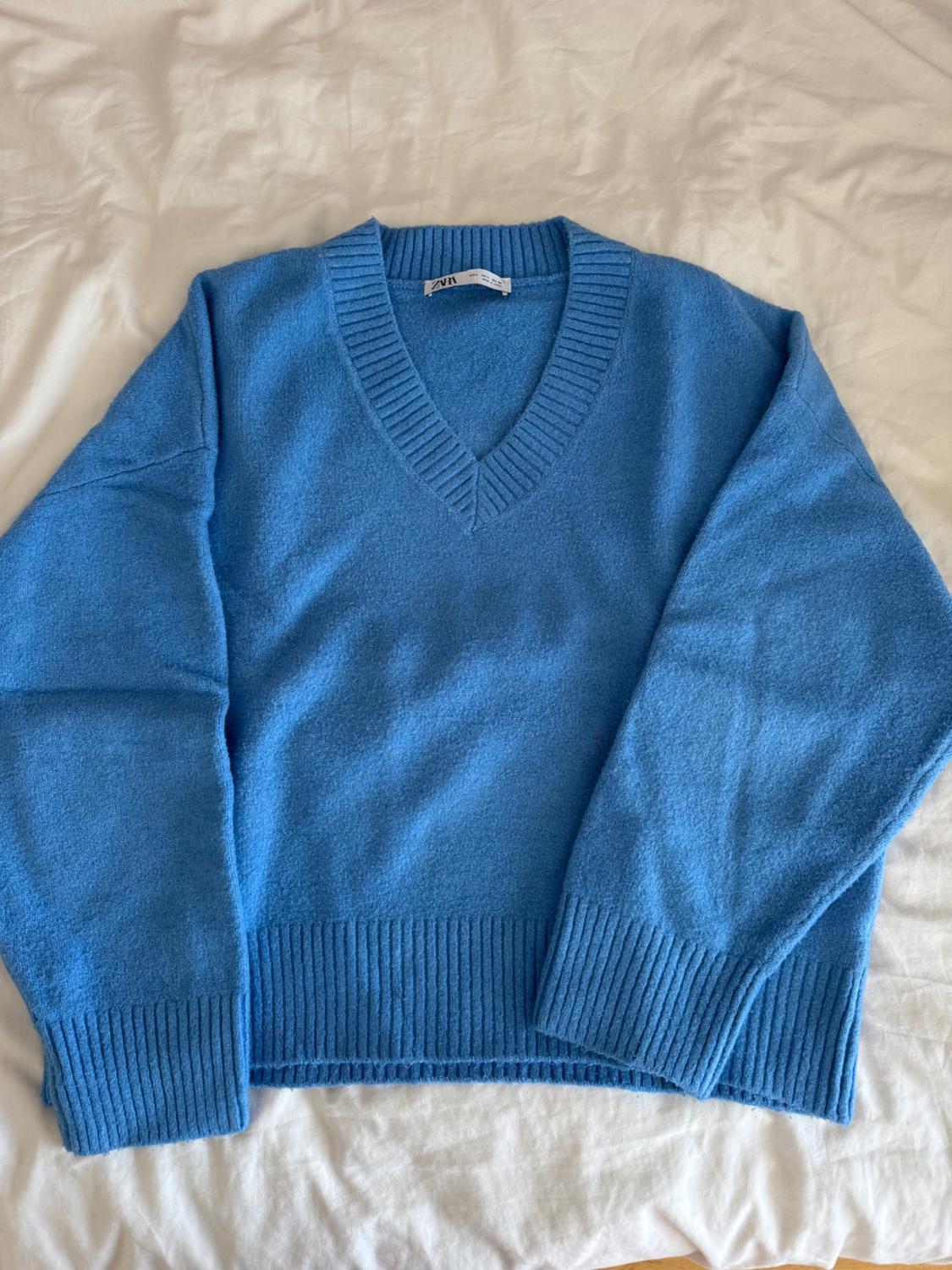Light blue knitted jumper, long sleeves and a V-neck, on a crumpled white sheet. The label reads ZARA.