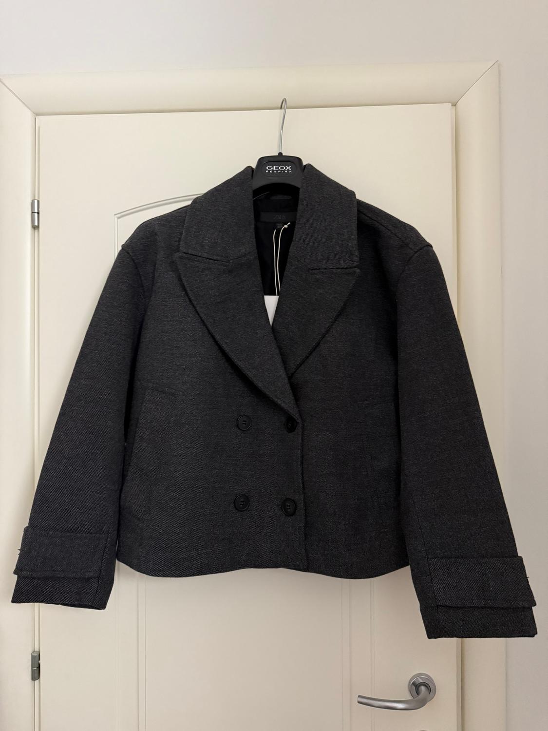 Dark grey double-breasted jacket hanging on a black branded hanger, over a white door.