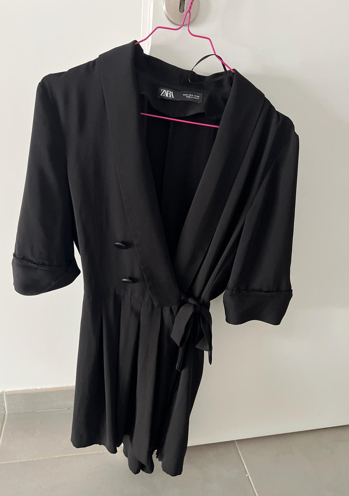 Black short-sleeved wrap dress with buttons and a tie, hanging on a pink hanger over a white door. Label with the text ZARA.